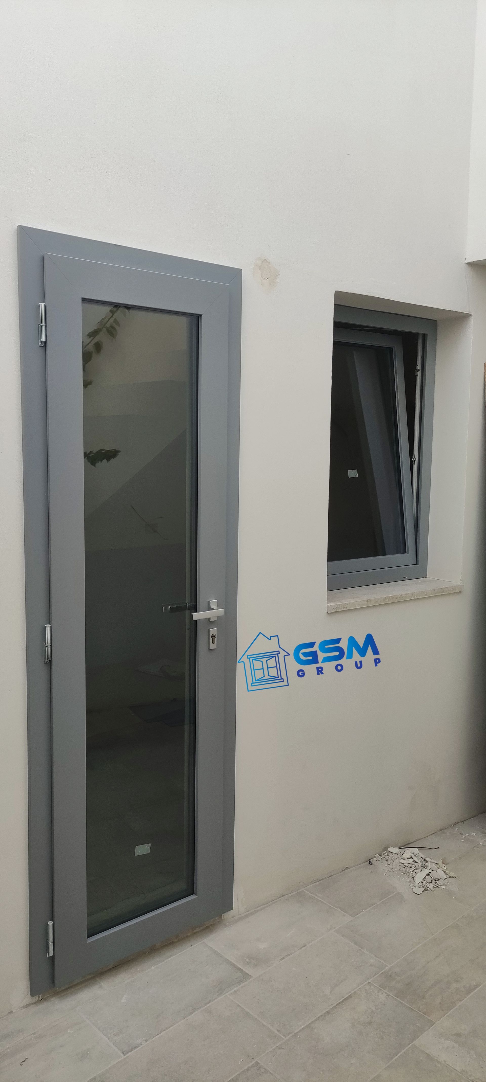 Una porta a vetri stretta con cornice grigia e una piccola finestra abbinata, apribile a vasistas, incastonate in una parete semplice di colore chiaro.