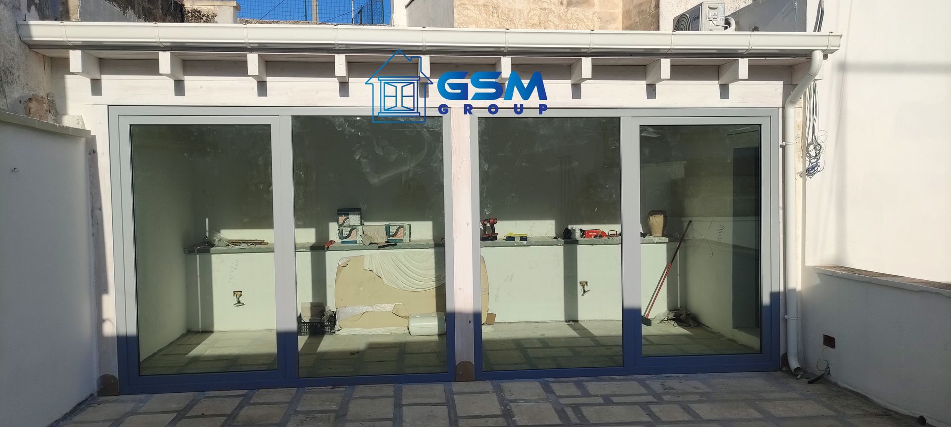 Un patio con un'ampia struttura a più pannelli di porte scorrevoli in vetro, situata sotto una pergola in legno bianco e decorata con il logo del Gruppo GSM.