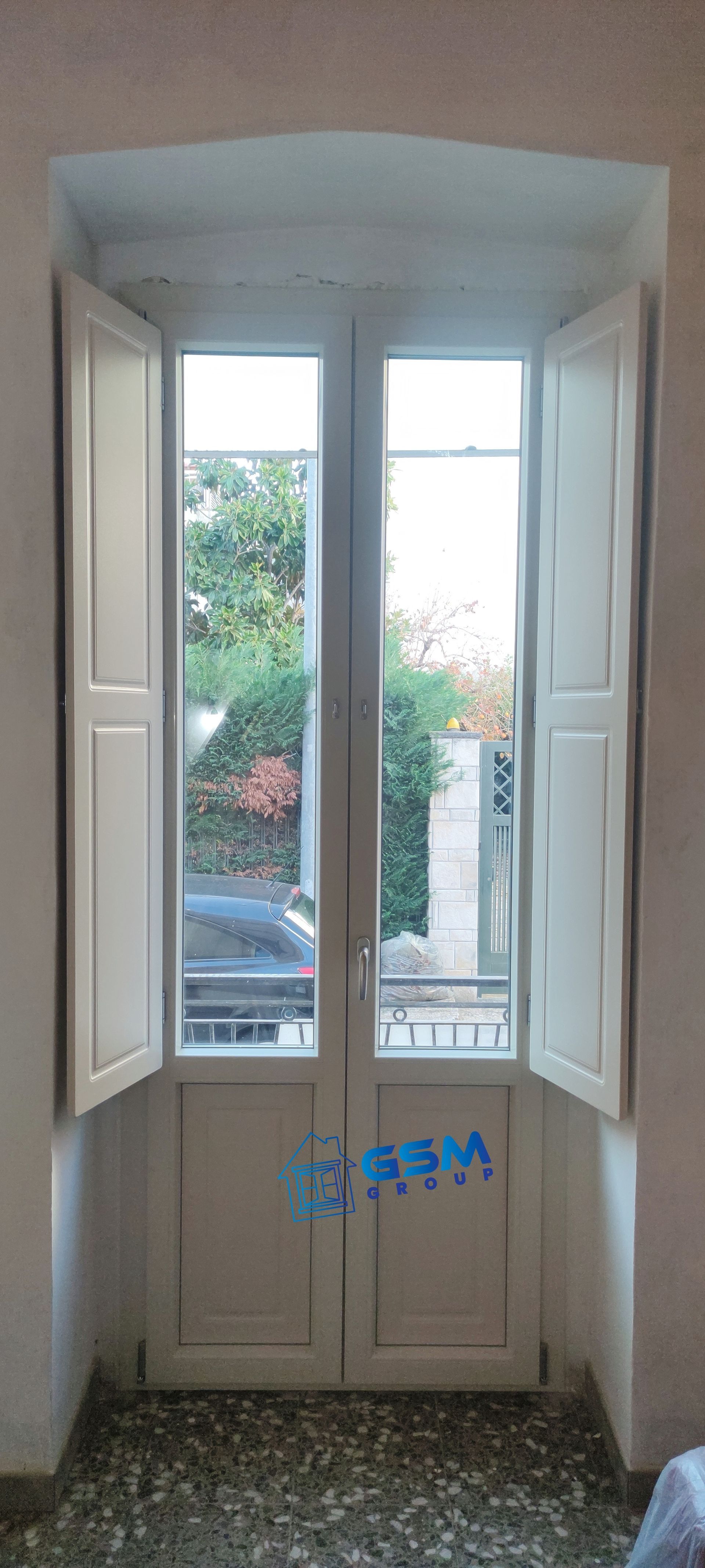 Una finestra alta e bianca a doppia anta con persiane aperte, incassata in una parete arretrata, che si affaccia su alberi e un veicolo parcheggiato.