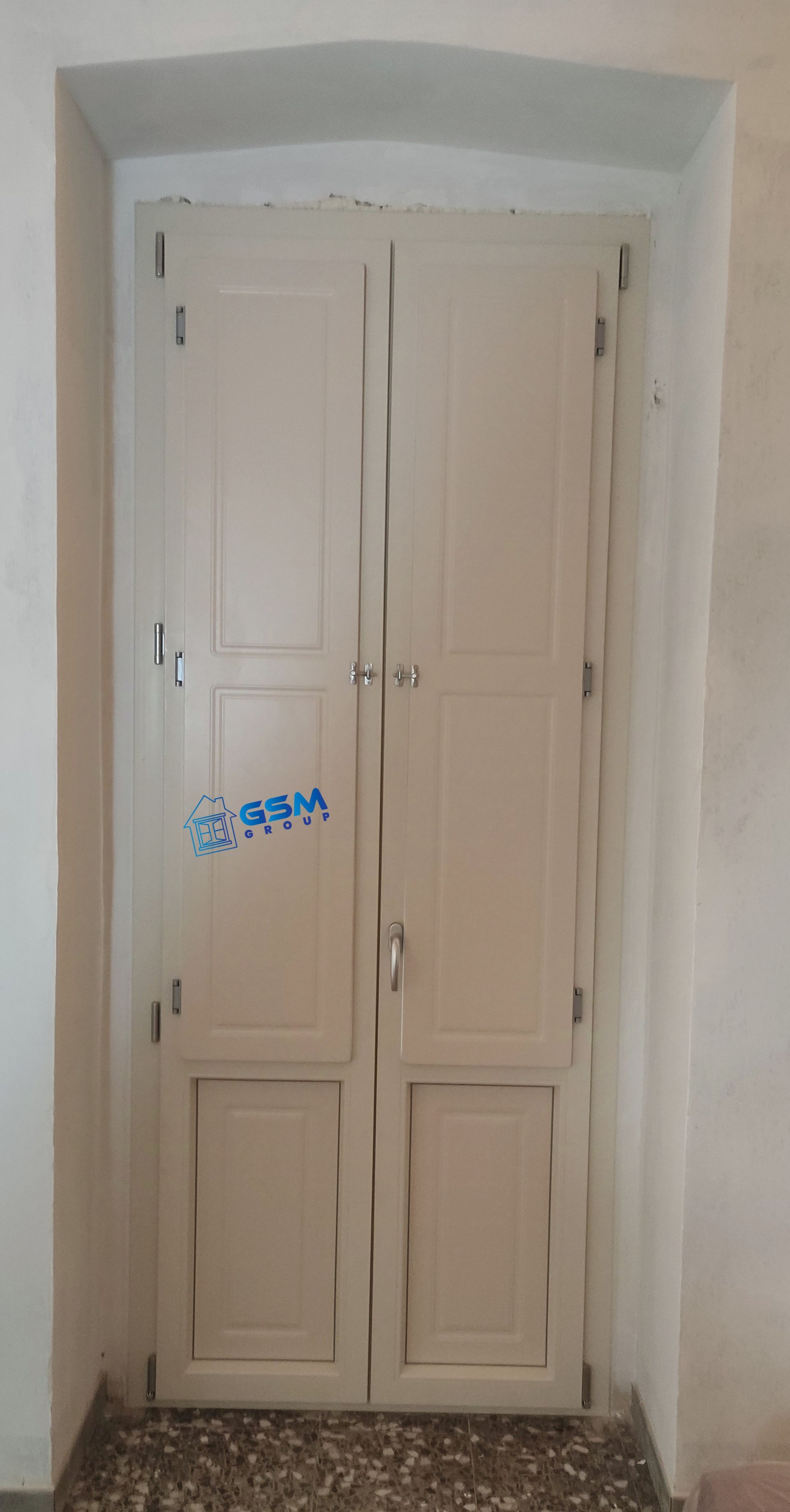 Una coppia di doppie porte in legno beige inserite in un profondo portale ad arco con pavimento screziato.