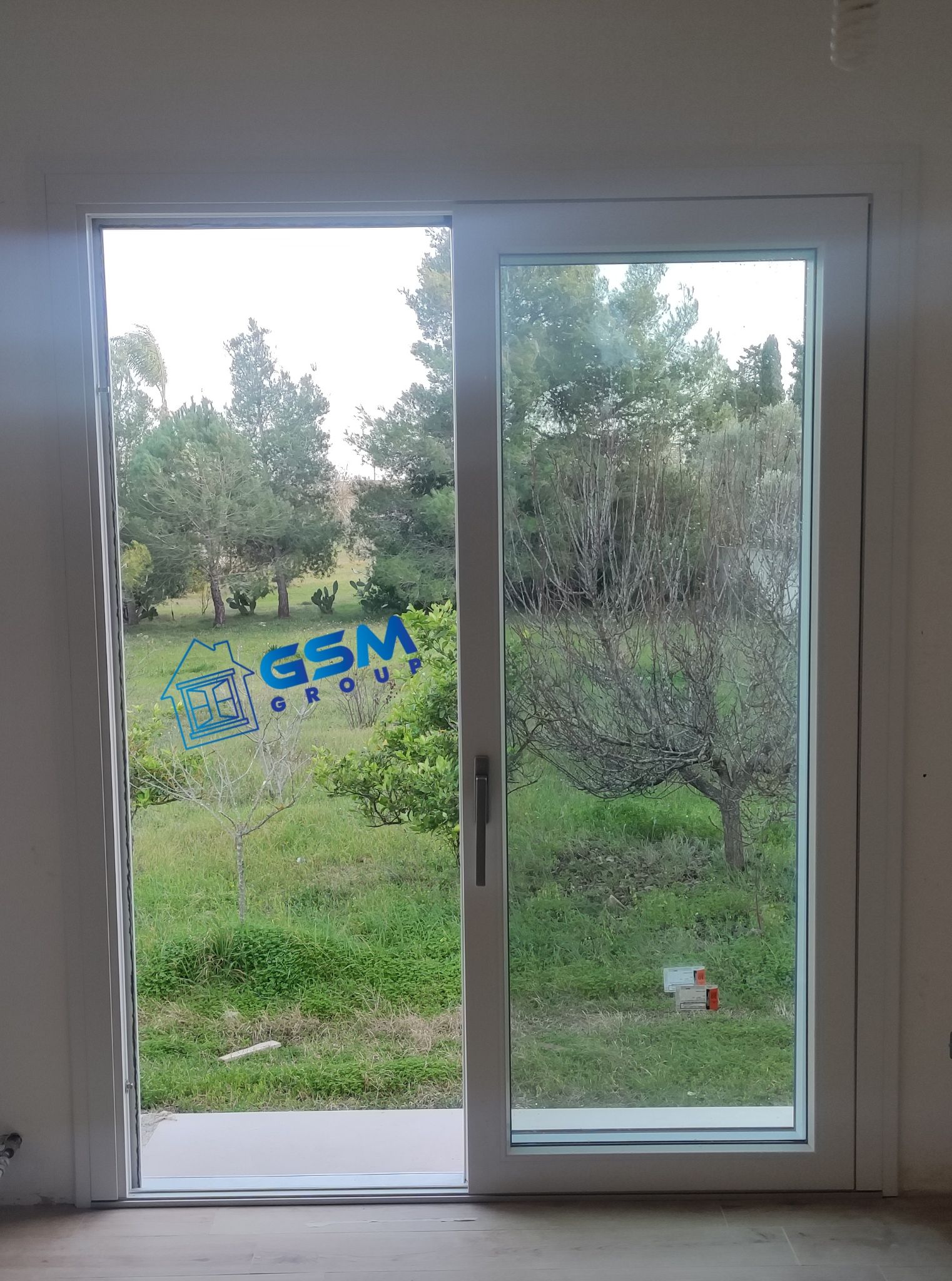 Porta scorrevole in vetro bianca che si affaccia su un prato alberato, con il logo blu del Gruppo GSM sul pannello di sinistra.