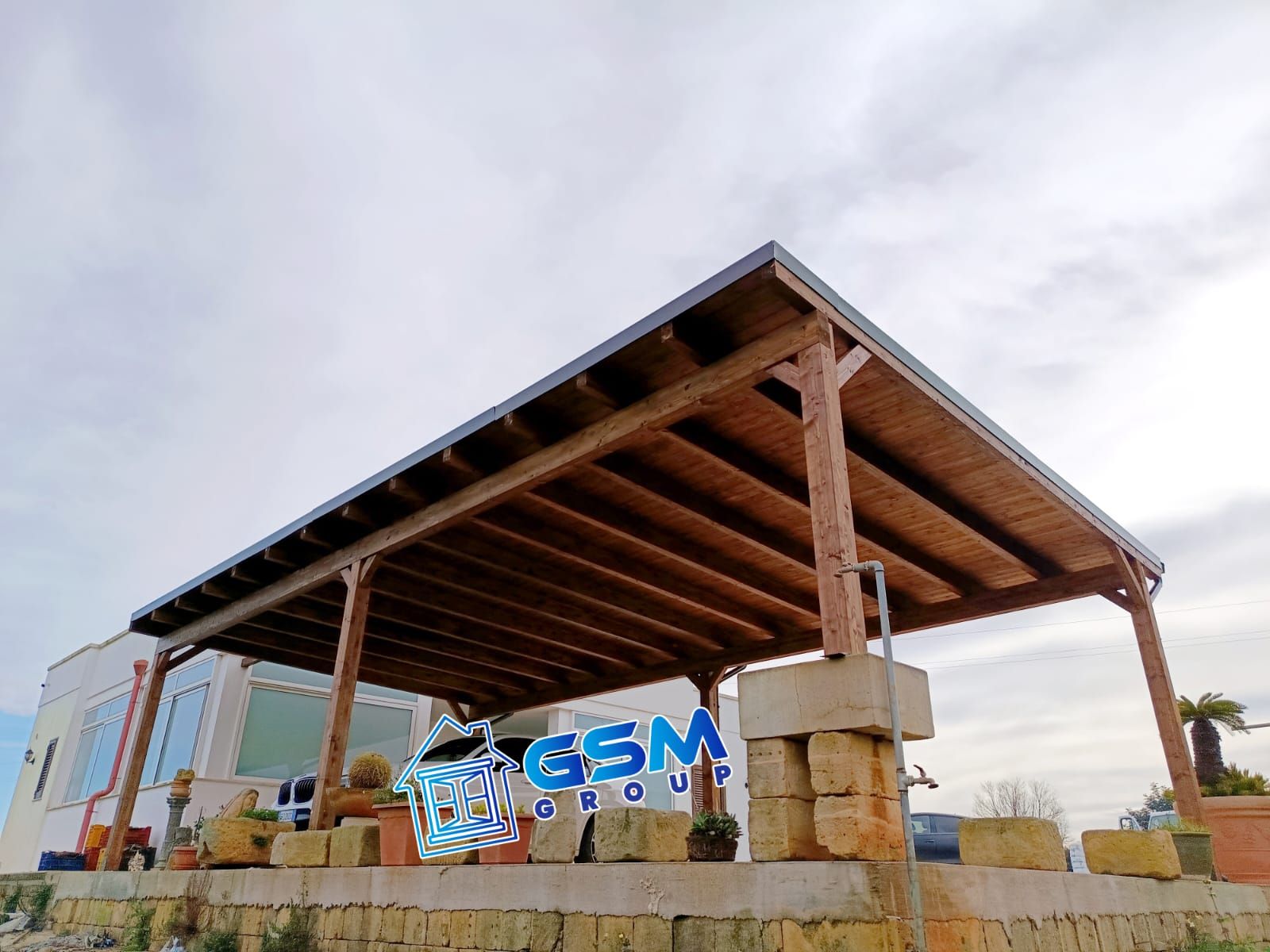 Una tettoia in legno per auto si erge di fronte a una casa moderna sotto un cielo nuvoloso, con un rubinetto dell'acqua in primo piano.