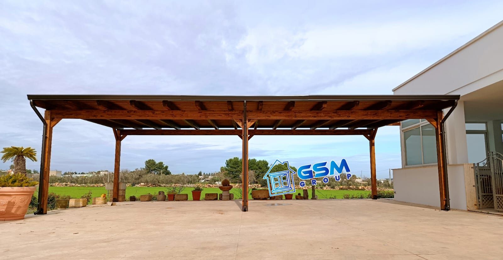 Una pergola in legno con tetto scuro si erge su un patio pavimentato accanto a un moderno edificio bianco, affacciata su un paesaggio verdeggiante.