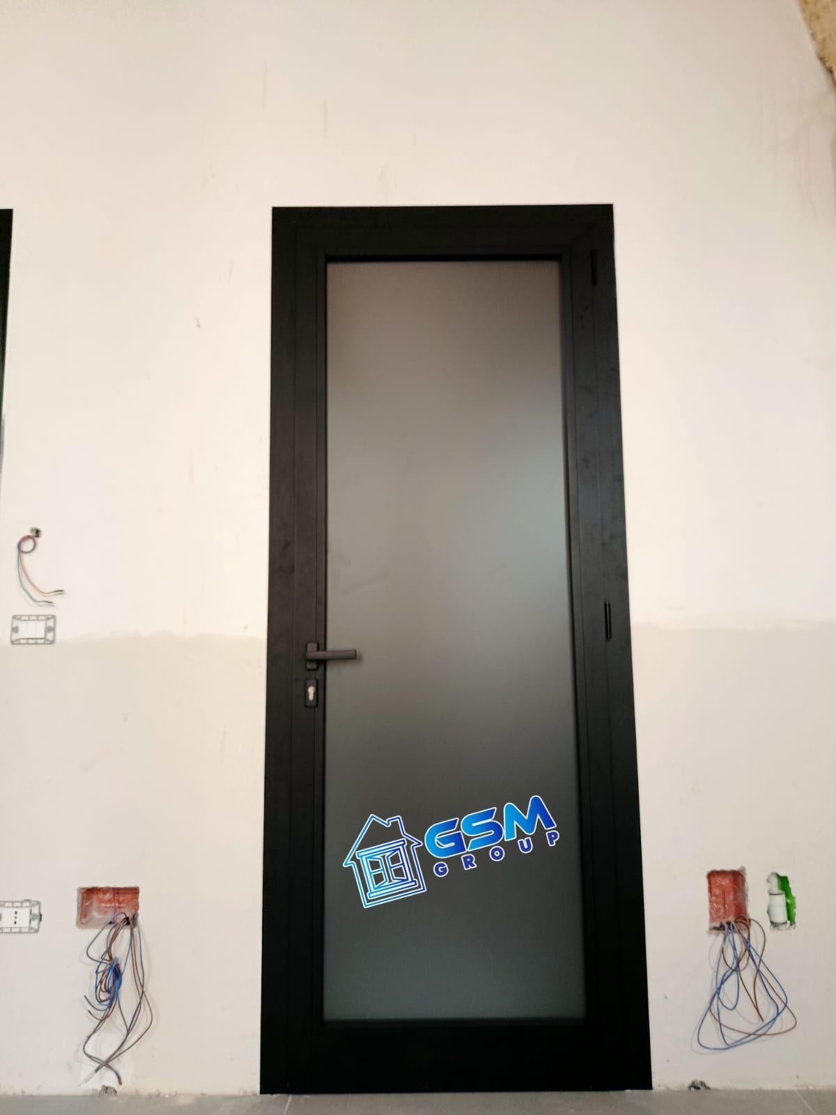 Una porta con cornice nera e pannello in vetro satinato, installata in una stanza non rifinita con impianto elettrico a vista.