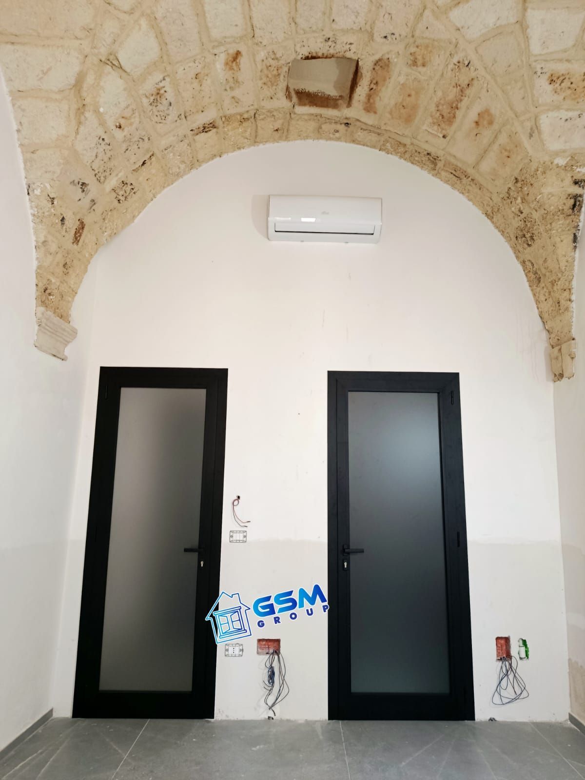 Una parete bianca con due porte nere sotto un arco in pietra, con un condizionatore d'aria a parete e i cavi a vista.
