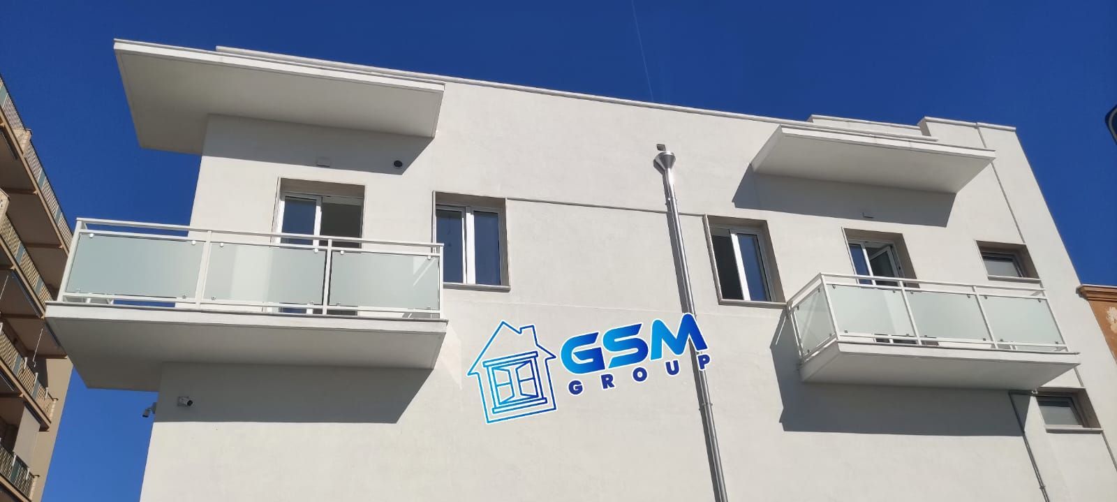 Vista dal basso di un moderno edificio di colore chiaro con balconi e un'insegna blu con la scritta 