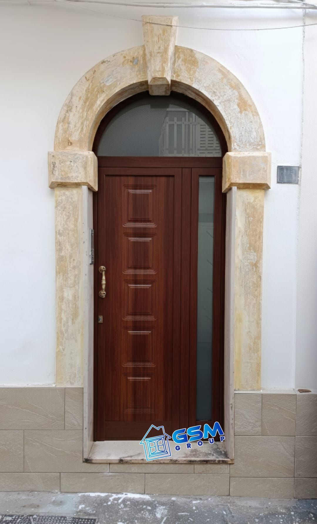 Una porta di legno marrone con un pannello laterale in vetro inserito in una cornice ad arco in pietra con una piccola finestra soprastante.