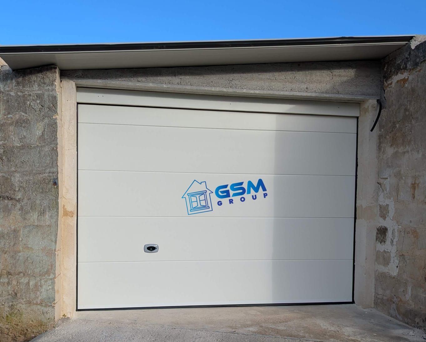 Una porta d'ingresso in legno con un pannello ad arco verticale, incastonata in una parete chiara con un piccolo citofono quadrato scuro.