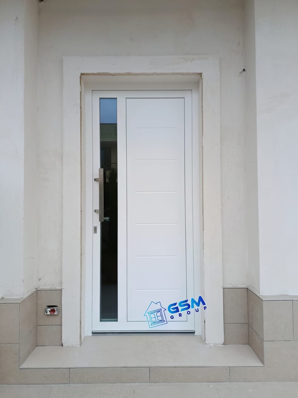 Una porta d'ingresso bianca con pannello verticale in vetro e maniglia lunga, installata in una parete esterna di colore chiaro.