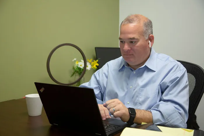 A person in a light blue shirt works on a laptop at a desk with a white mug and decorative ring in a simple office.