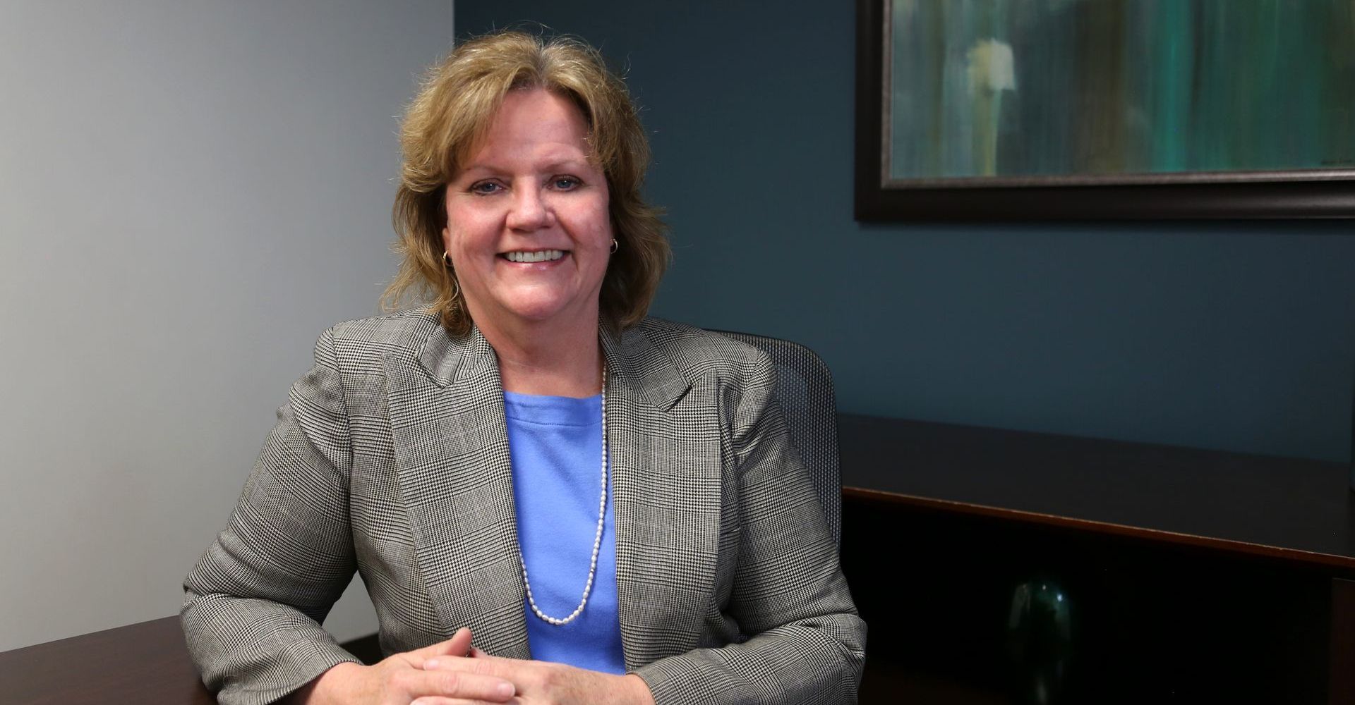 A woman is sitting at a desk with her hands folded and smiling.