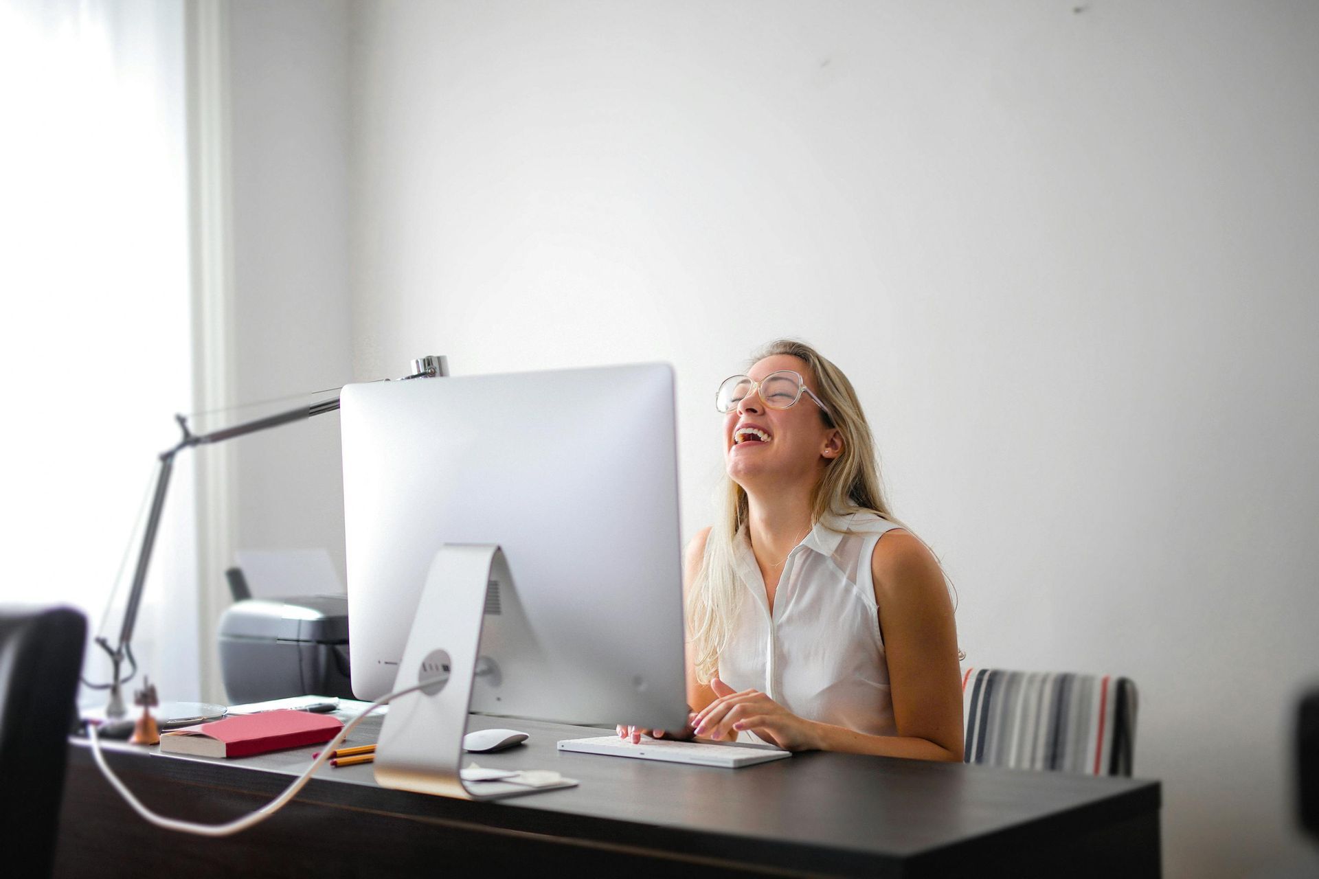 A person sitting at a desk with a computer monitor, laughing while looking up with their mouth open.