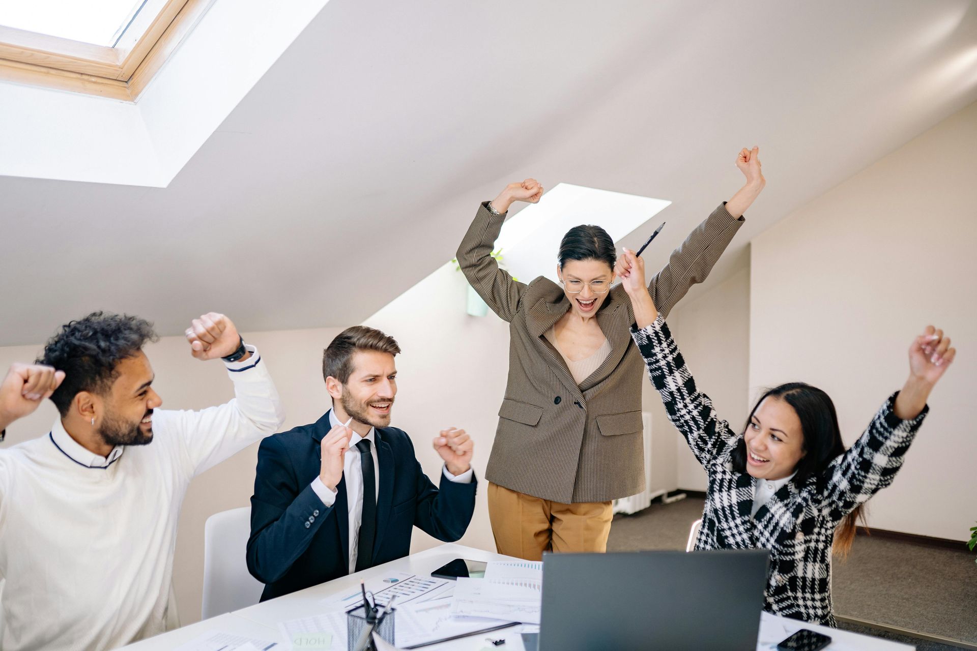 Four colleagues in an office celebrating success with their arms raised while looking at a laptop on their desk.