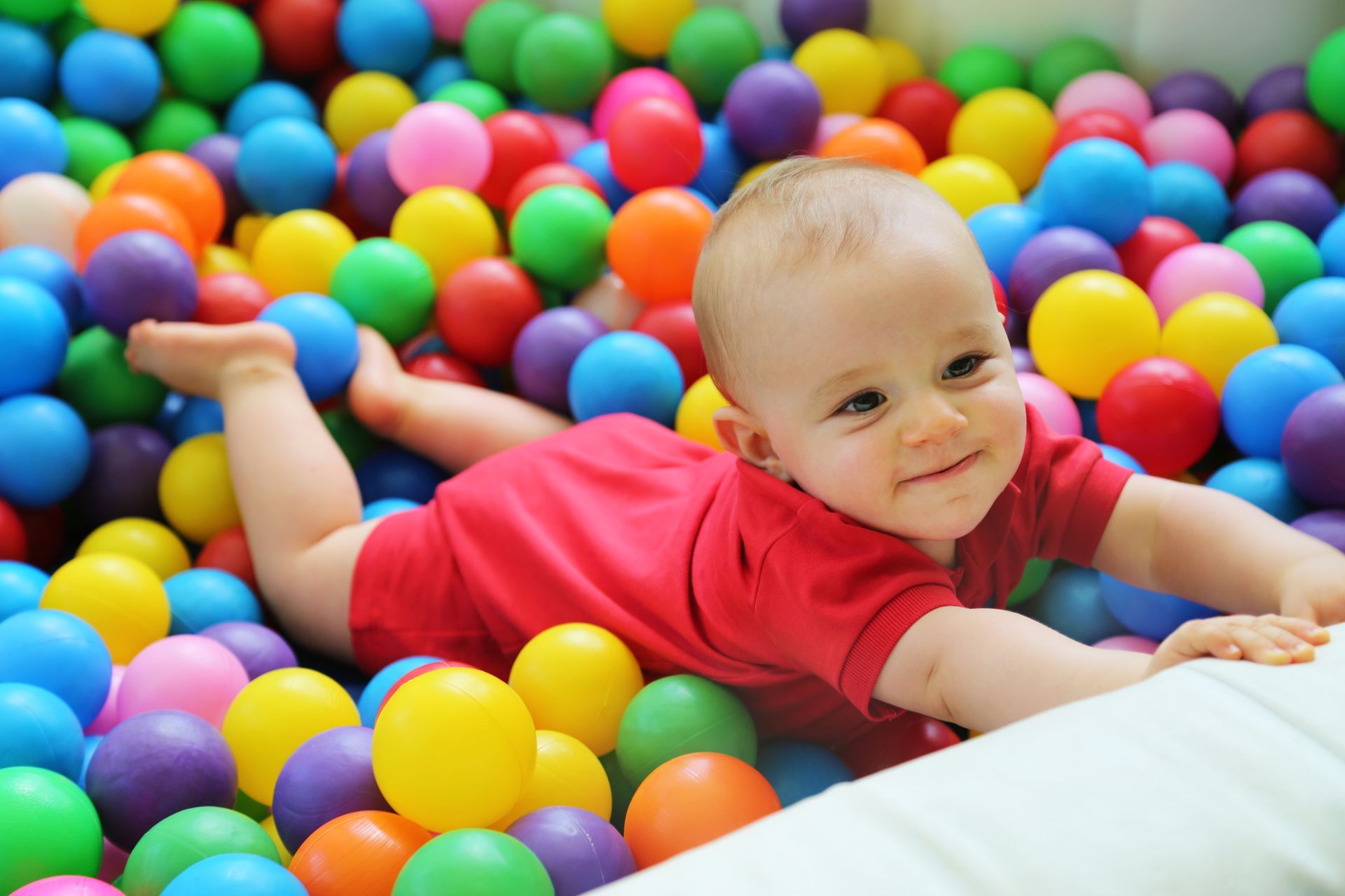 Baby in red onesie smiles while crawling through colorful ball pit.