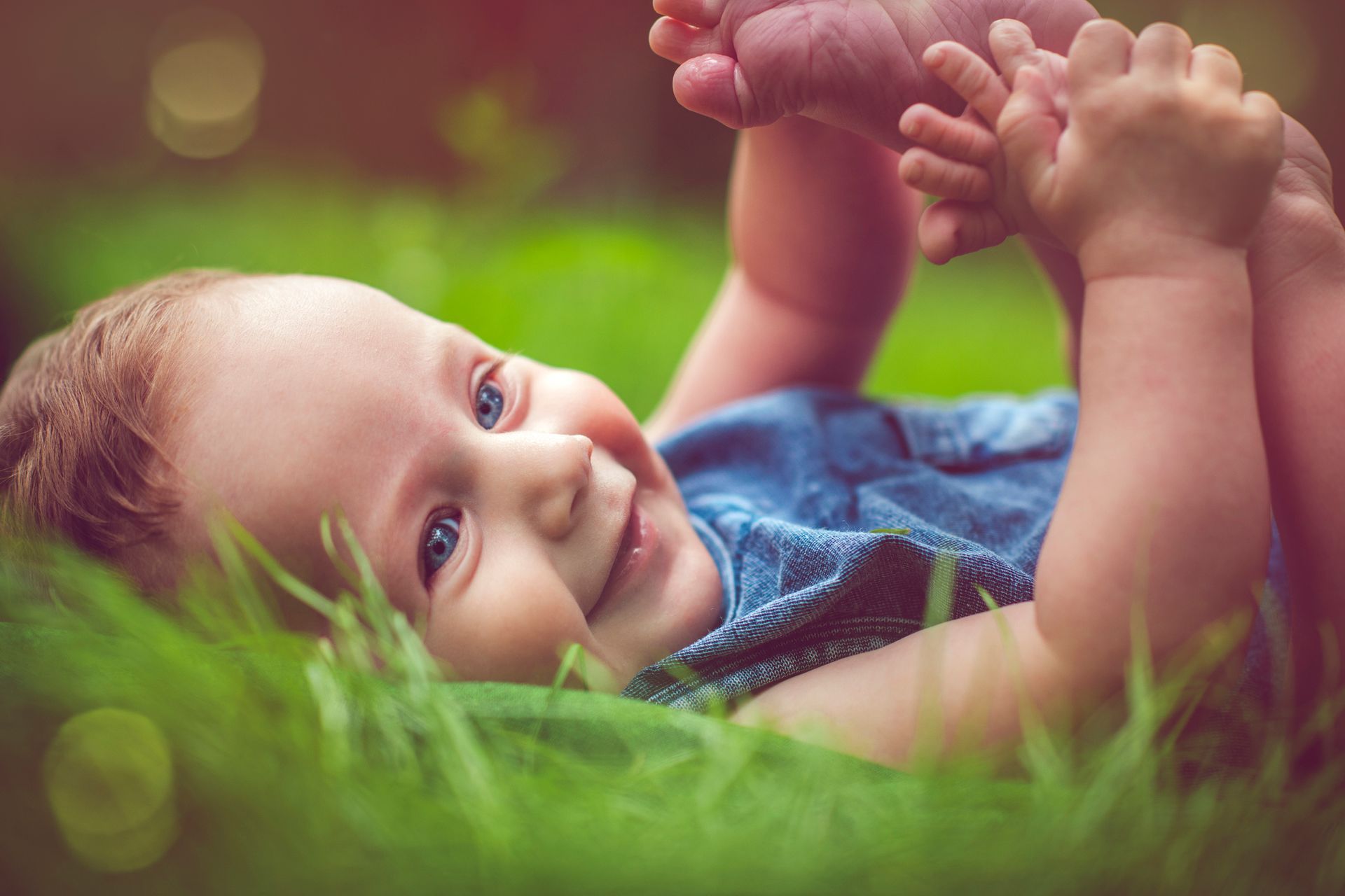 Smiling baby in denim lying in green grass, reaching for toes.