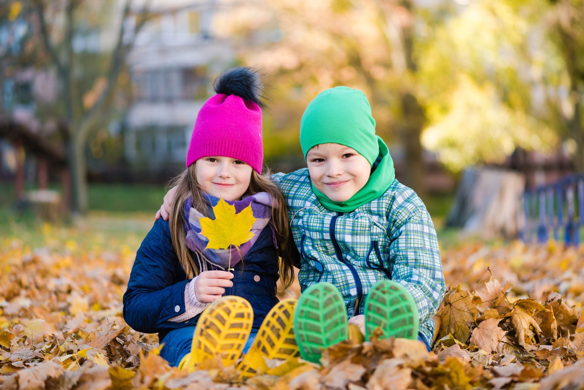 Two children in beanies smiling, surrounded by autumn leaves, one holding a leaf.