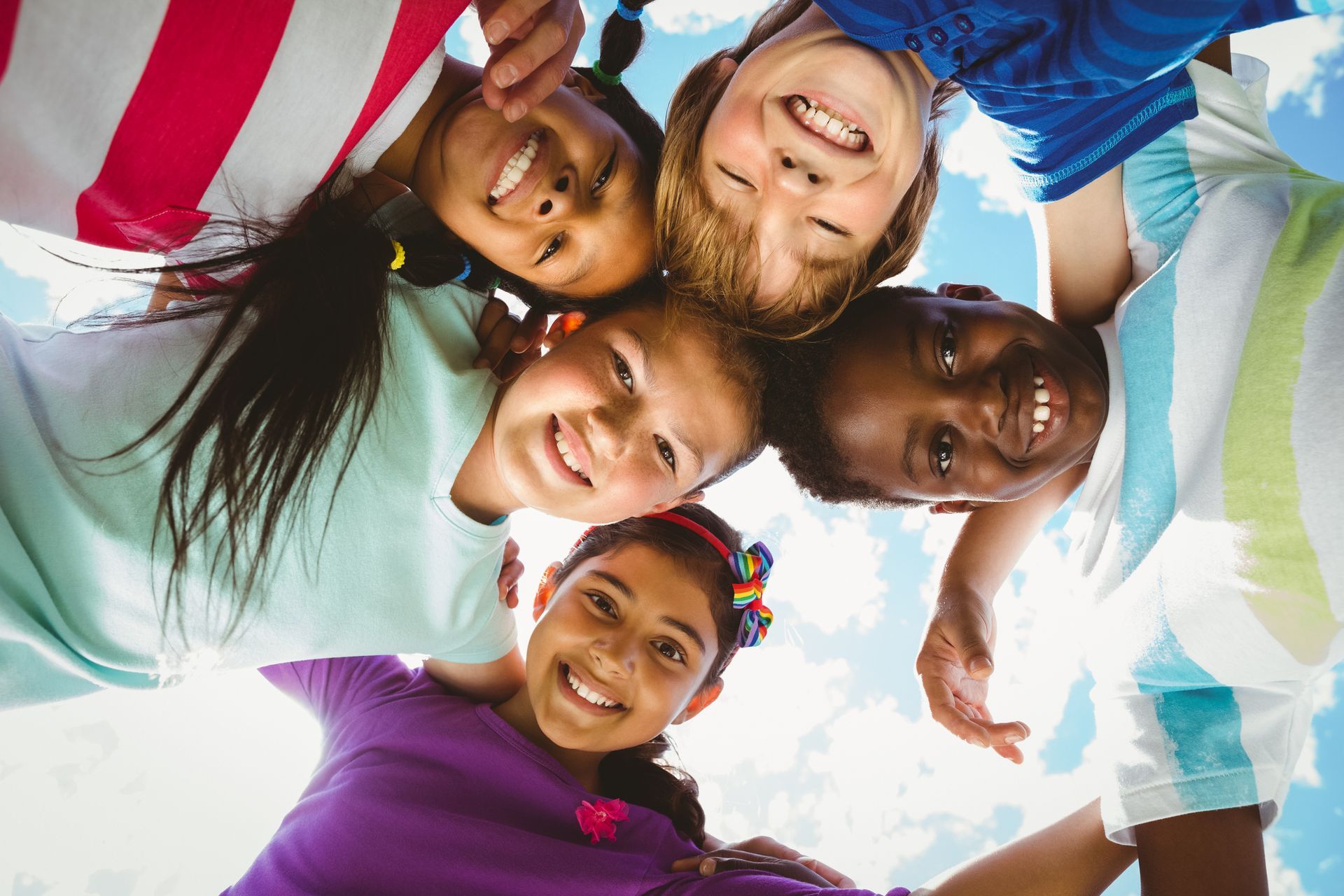 Children smiling, looking down, gathered closely together, blue sky in background.