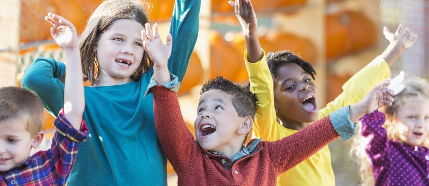 Children with arms raised, smiling and excited outdoors, orange pumpkins in the background.