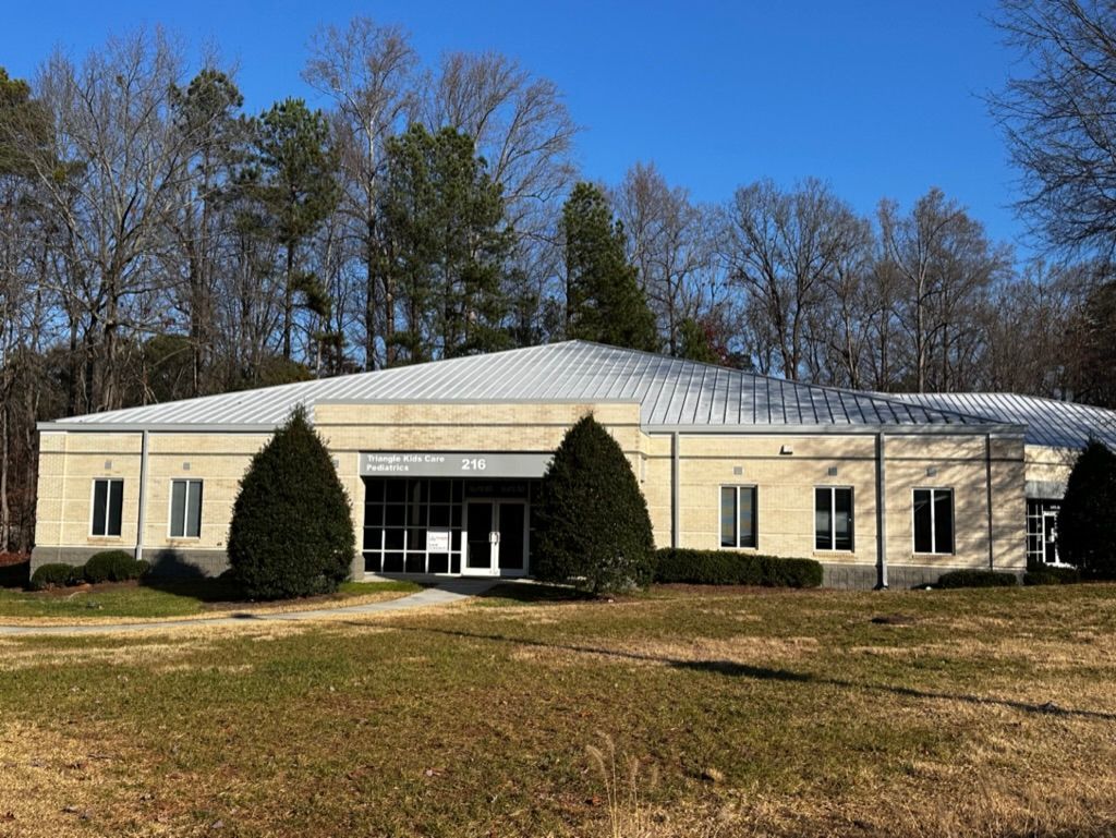 Cream-colored commercial building with a silver roof, windows, and manicured shrubs in front. Trees in the background.
