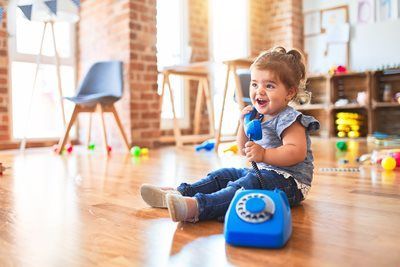 Smiling toddler sits on the floor, holding a blue toy telephone in a brightly lit room.