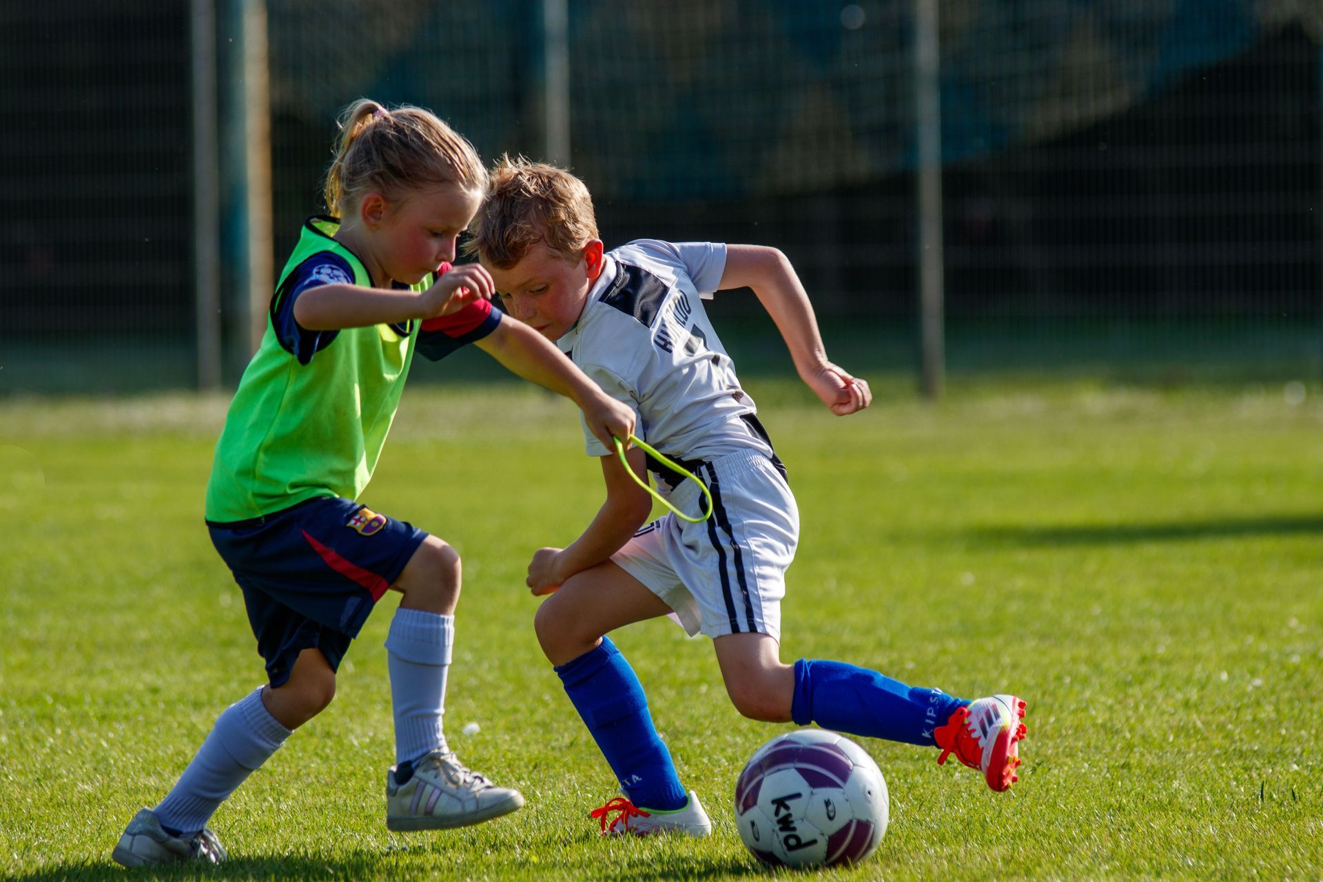 Two children playing soccer on a sunny field, chasing a ball near the goal