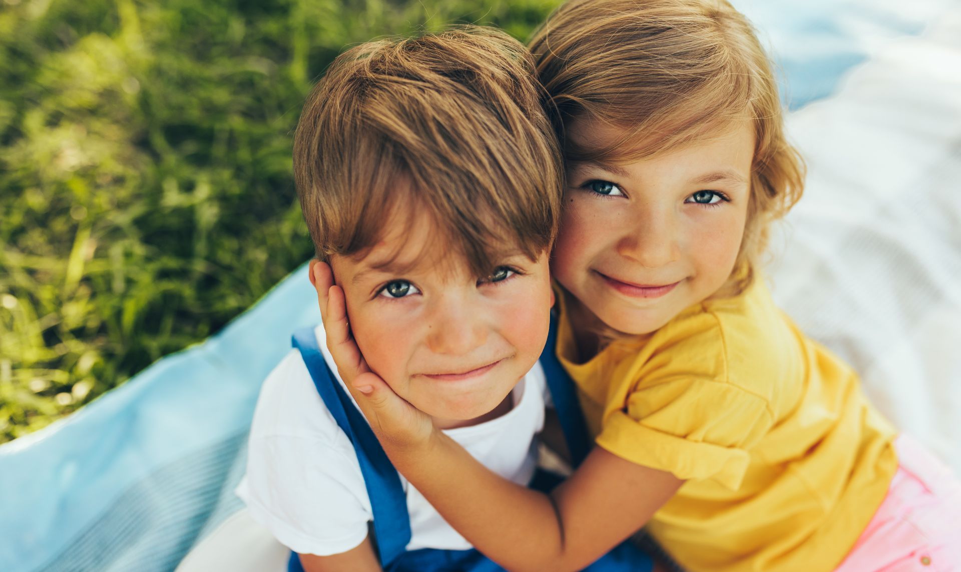 A girl embraces a boy, both smiling. They sit on a blue blanket on grass.