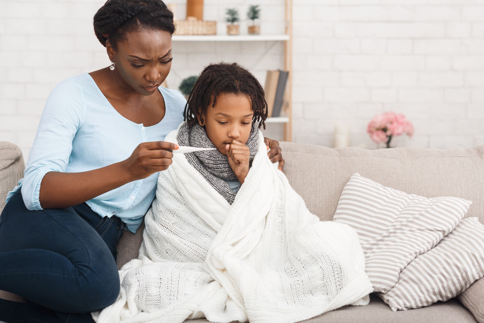 Woman taking a child's temperature on a sofa; the child is wrapped in a blanket, looking unwell.