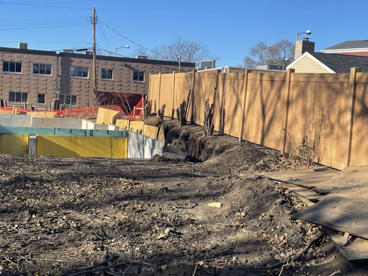 A wooden fence surrounds a construction site with a brick building in the background