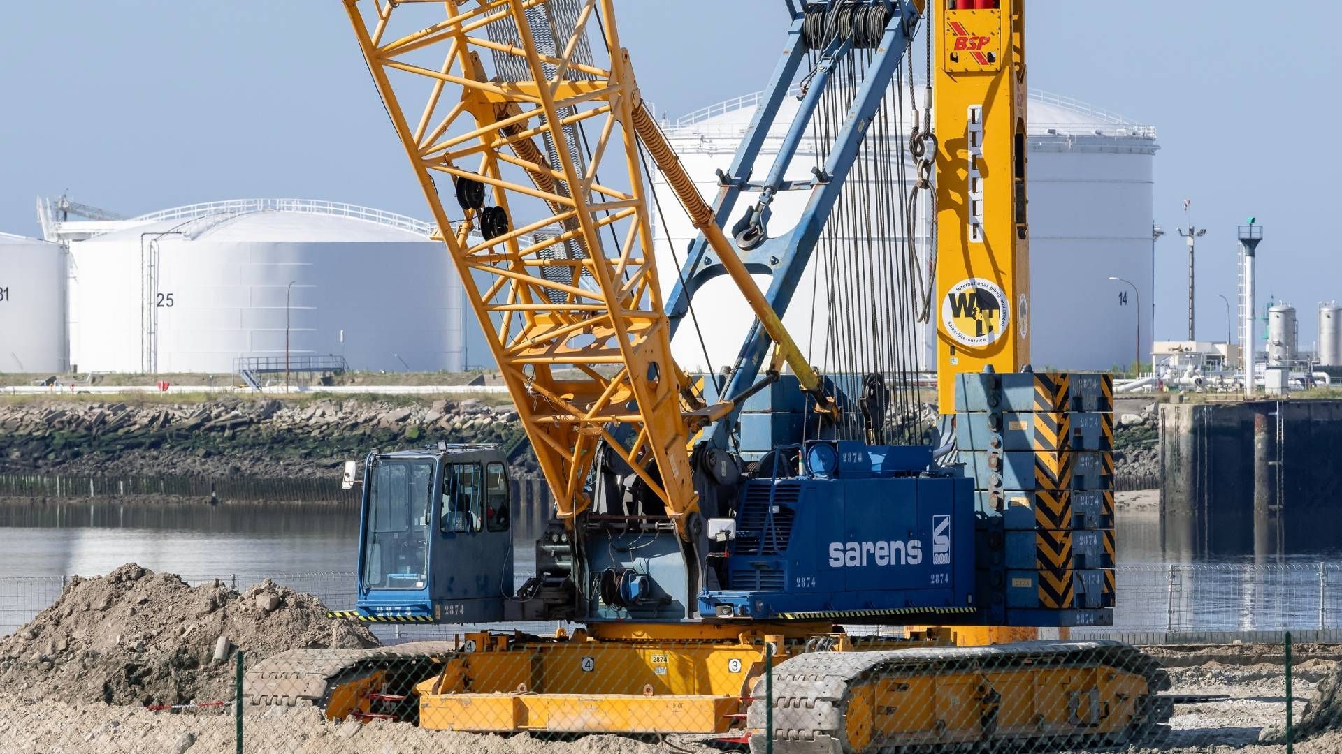 Yellow and blue crane on tracks at a construction site with white storage tanks in the background.