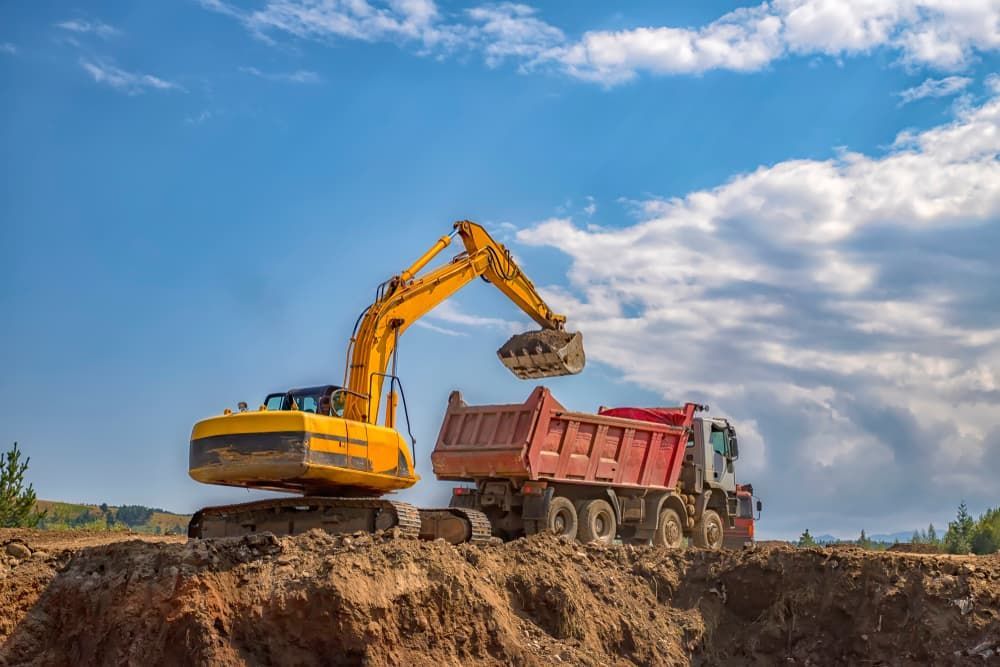 Yellow excavator loading a red dump truck with dirt on a construction site under a blue sky.