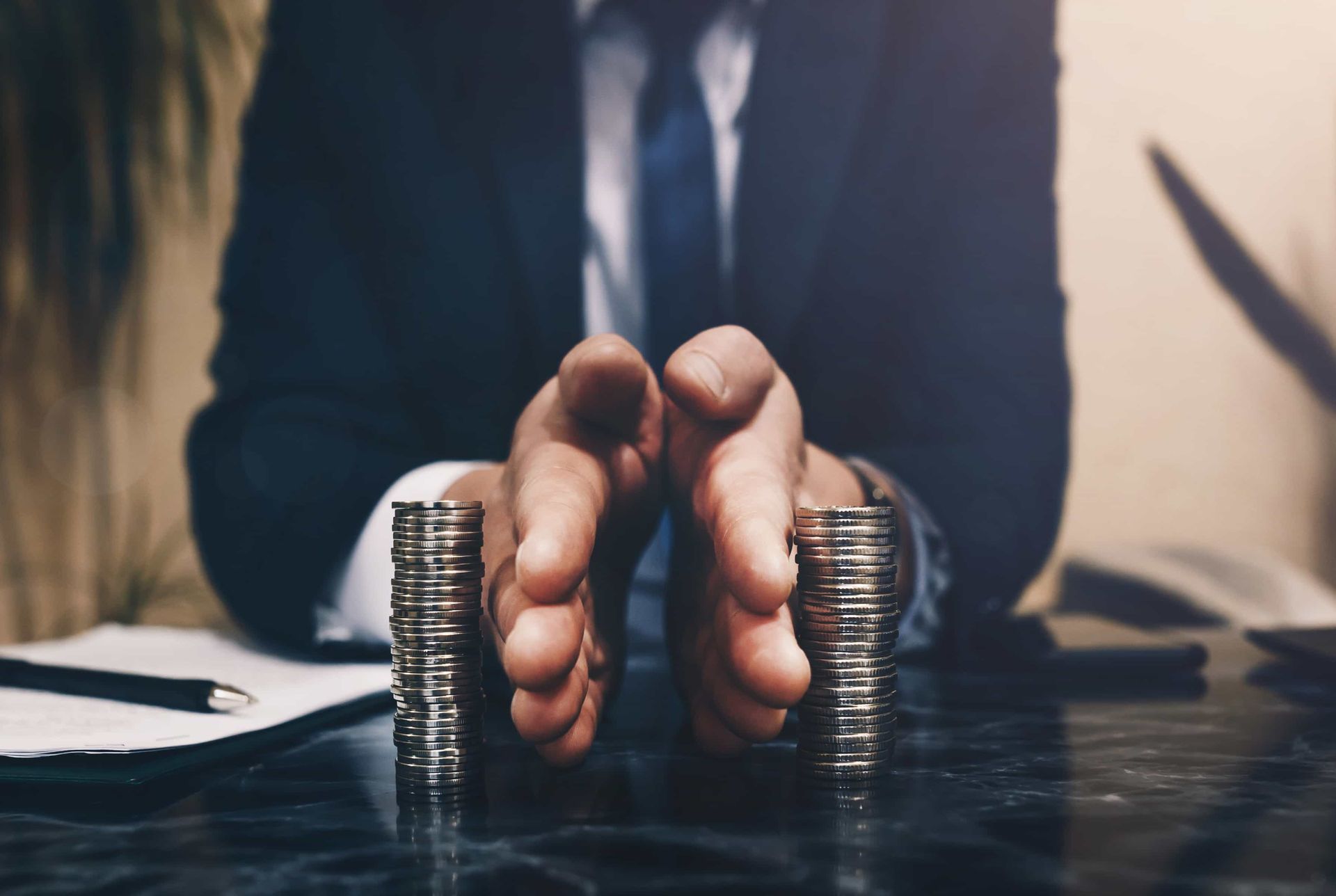 Person in suit protects stacks of coins with their hands on a dark surface.