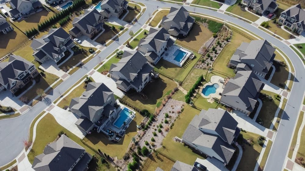 Aerial view of suburban houses with gray roofs, green lawns, and curving streets. Some have pools.