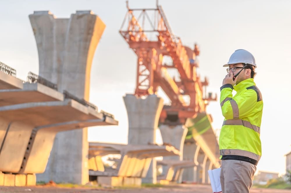 Construction worker in yellow vest and hard hat using a walkie-talkie at a bridge construction site.