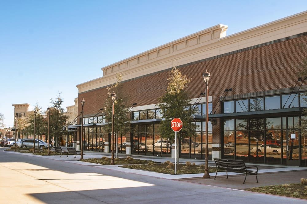 Street with shops: brick buildings, glass storefronts, benches, stop sign, trees, clear sky.