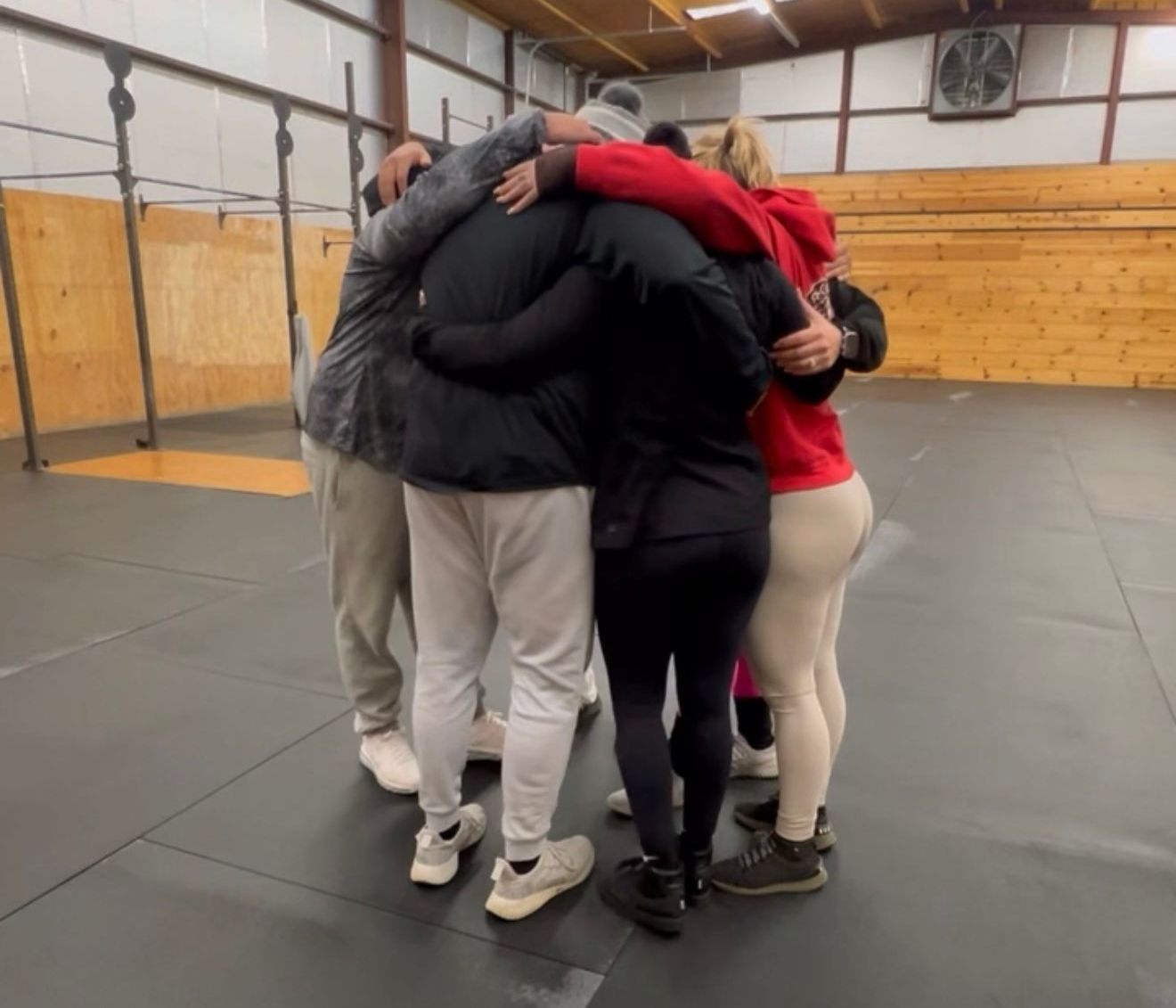 A group of people in athletic wear huddle together, arms around each other, in a gym.