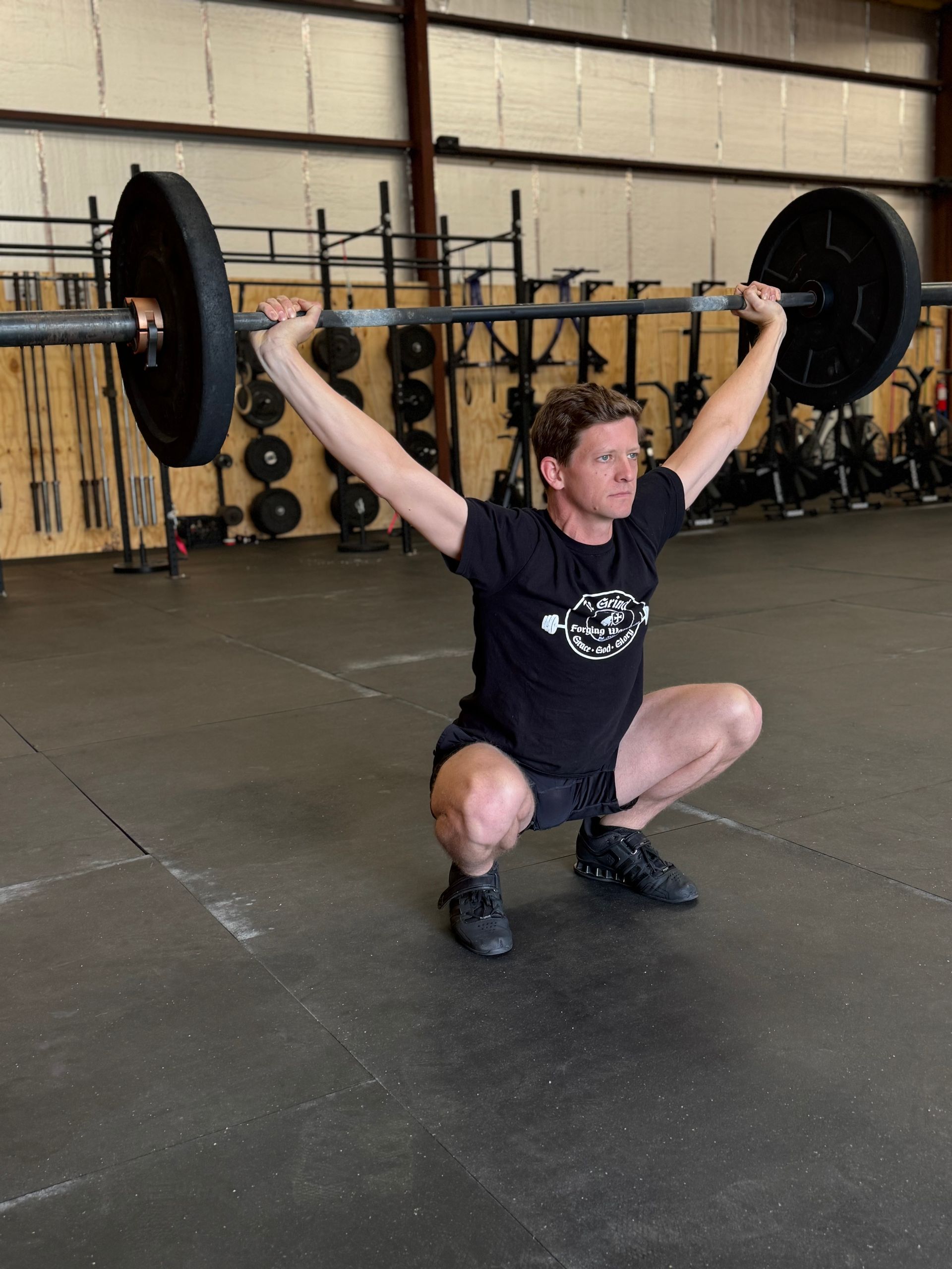 A man in a black shirt performs an overhead squat with a barbell in a gym. He’s in a low squat with arms fully extended.
