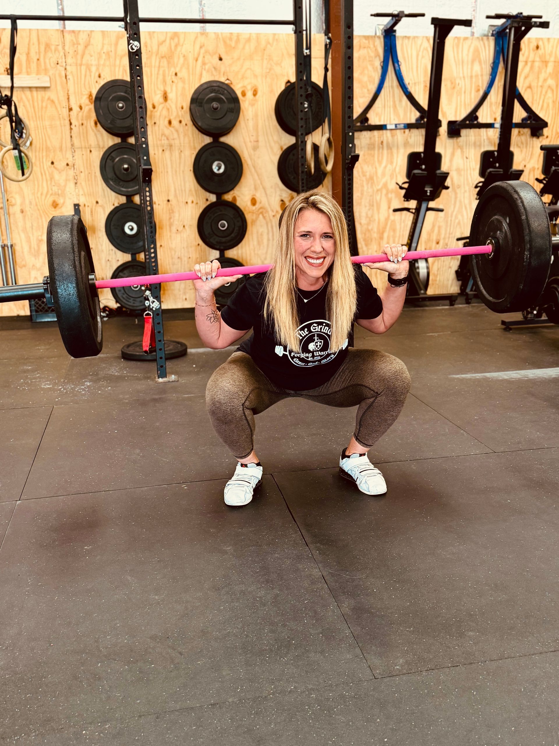 Woman with blonde curly hair smiles while squatting with a barbell in a gym.