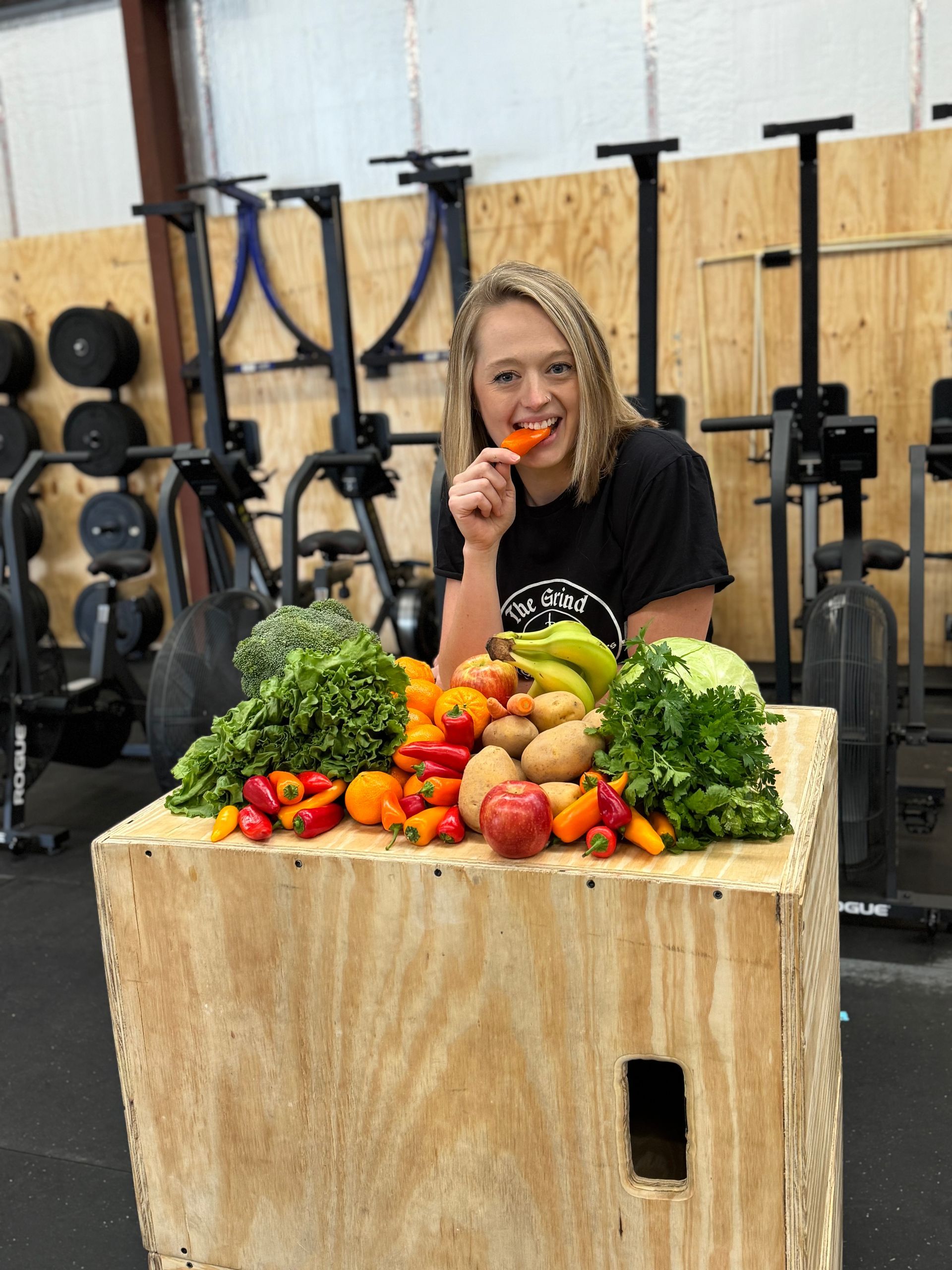 Woman biting a pepper, surrounded by colorful produce on a wooden box in a gym setting.