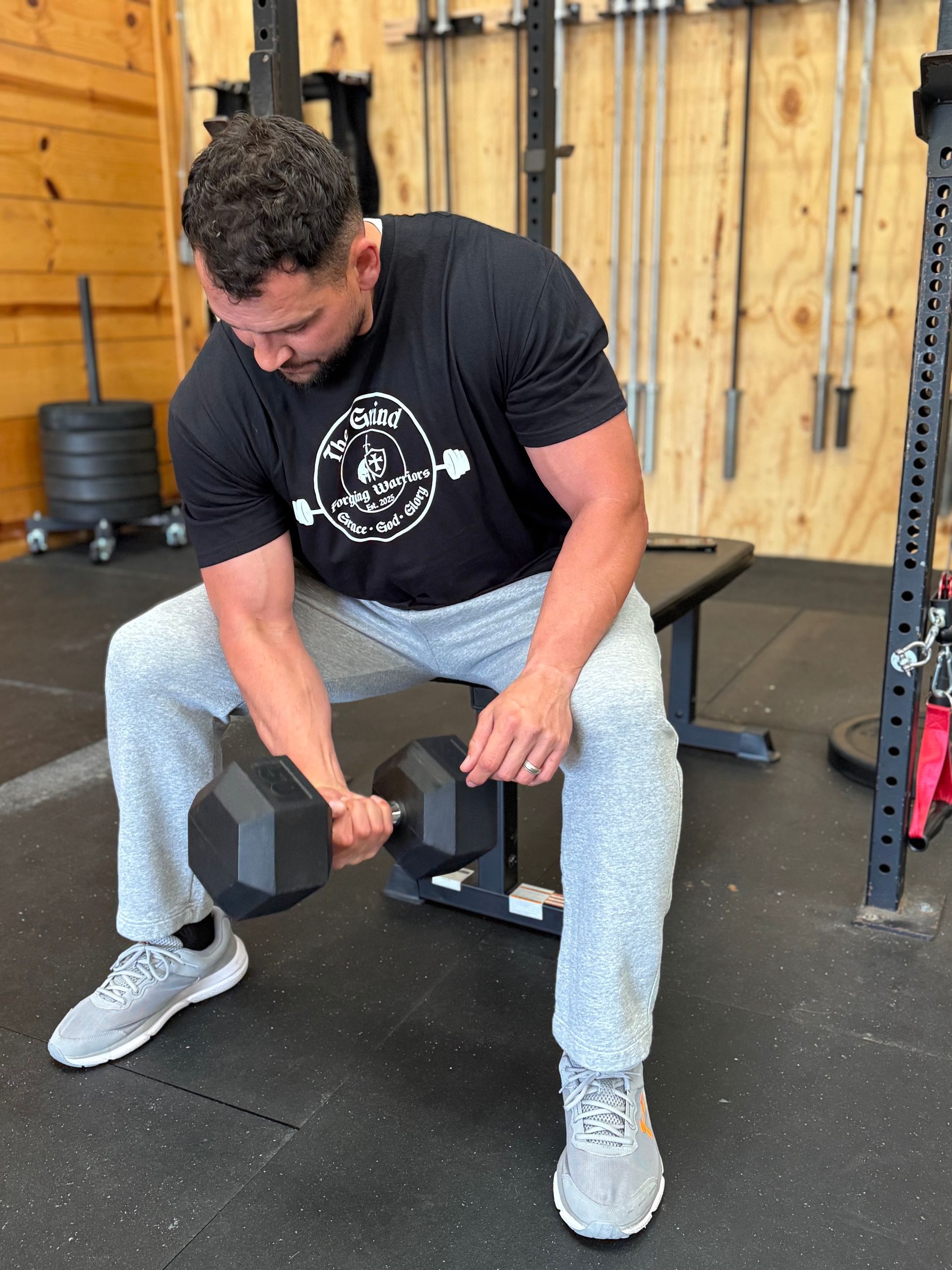 Man in a gym doing dumbbell bicep curls while seated on a bench. He is wearing gray sweatpants and a black t-shirt.