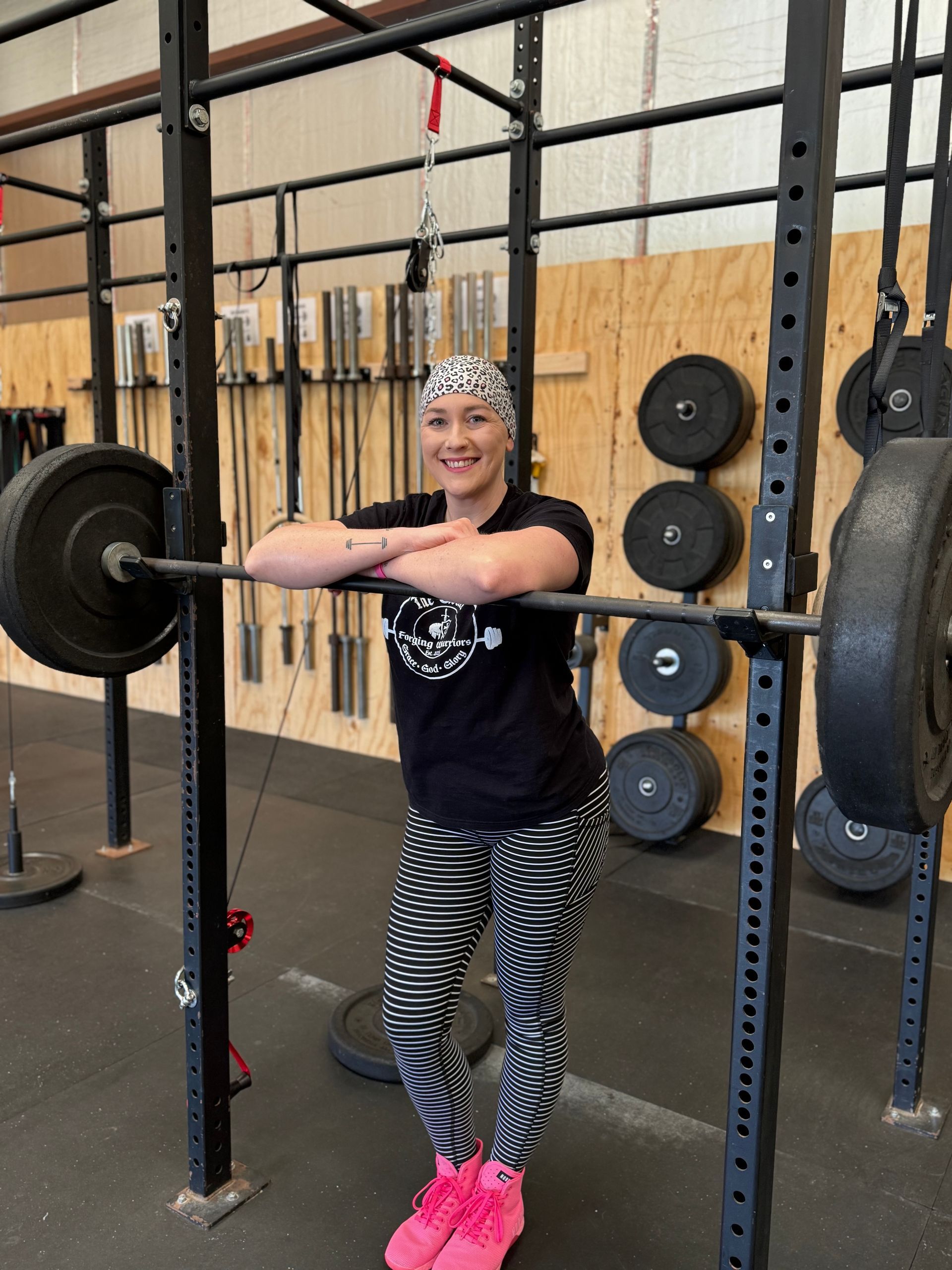 Woman in a gym leaning on a barbell, smiling. She wears a headscarf, black shirt, patterned leggings, and pink shoes.