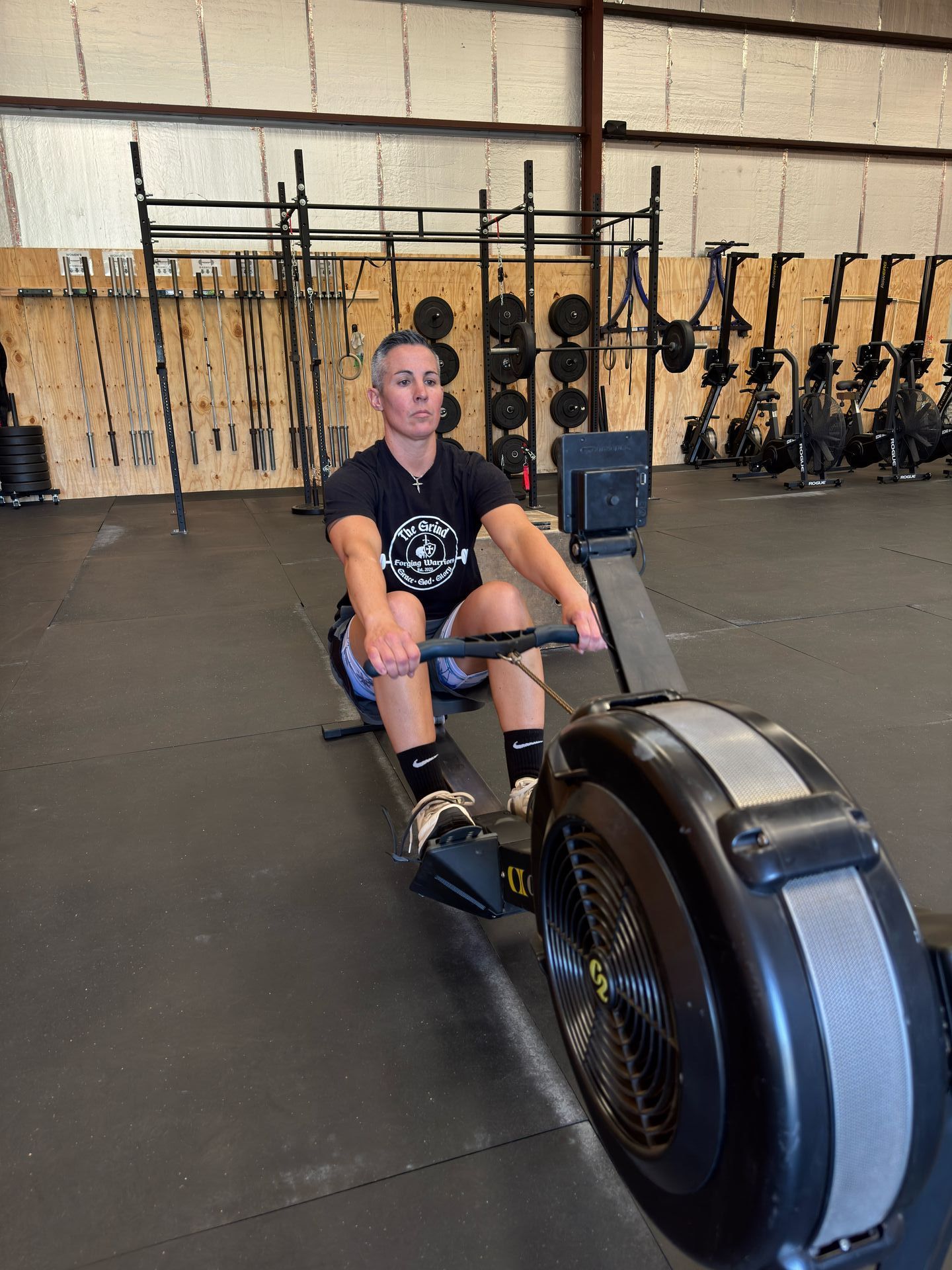 A person with short grey hair is rowing on a stationary machine in a gym. They are wearing a black shirt and shorts, and appear focused.