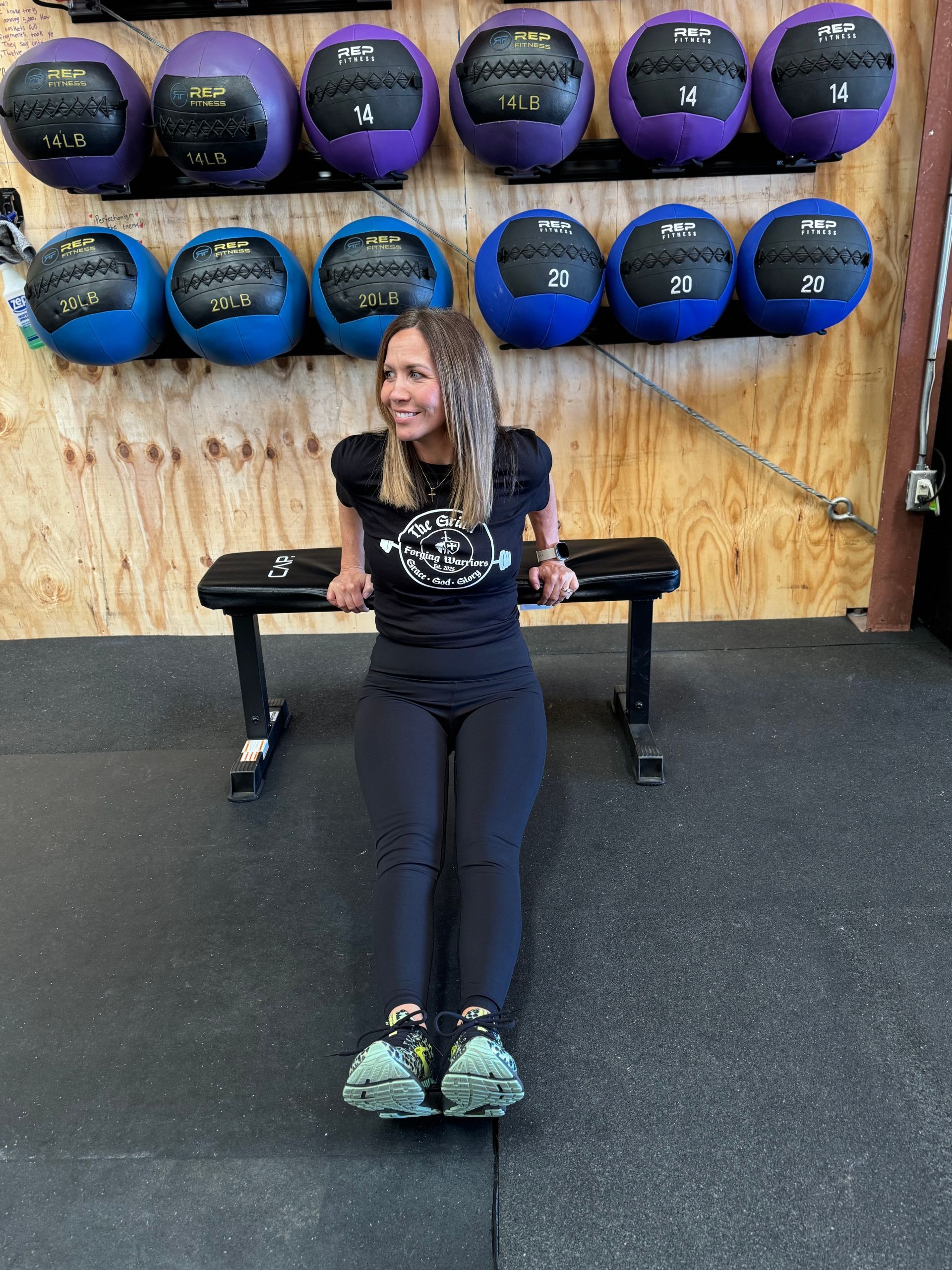 Woman doing tricep dips on a bench in a gym. She wears black athletic wear and smiles while exercising. Medicine balls are mounted on the wall behind her.