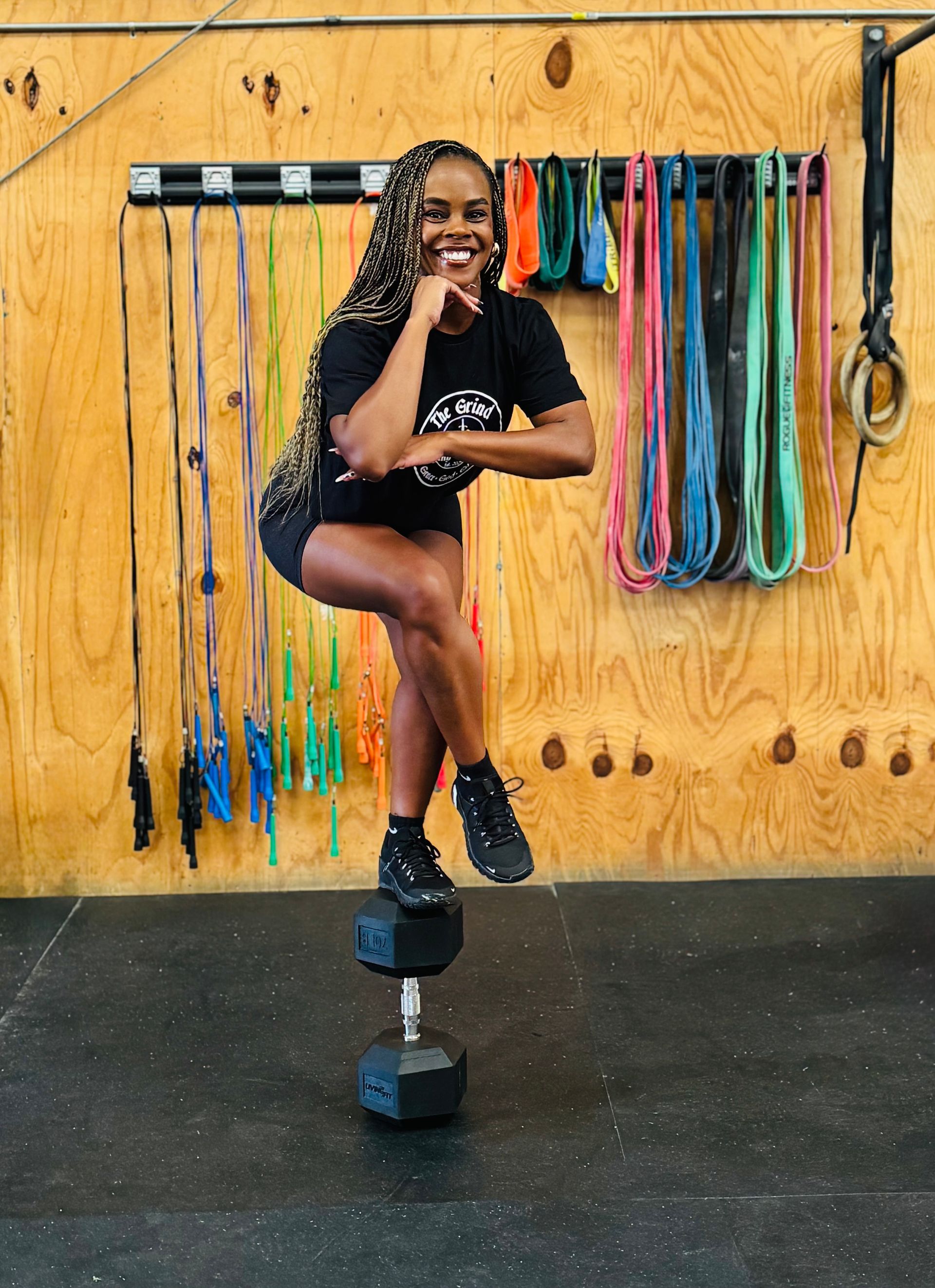 A woman balances on a dumbbell in a gym, smiling with one hand on her chin. She's wearing black athletic clothes. Jump ropes hang in the background.