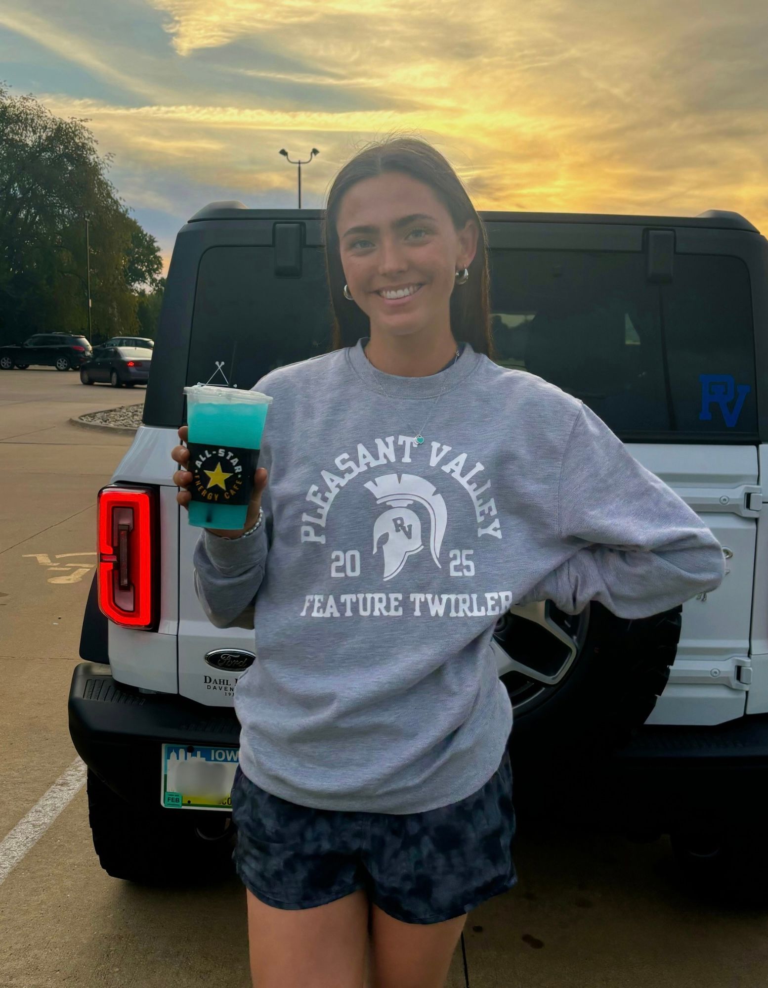 A woman in a pleasant valley sweatshirt is holding a drink in front of a jeep.