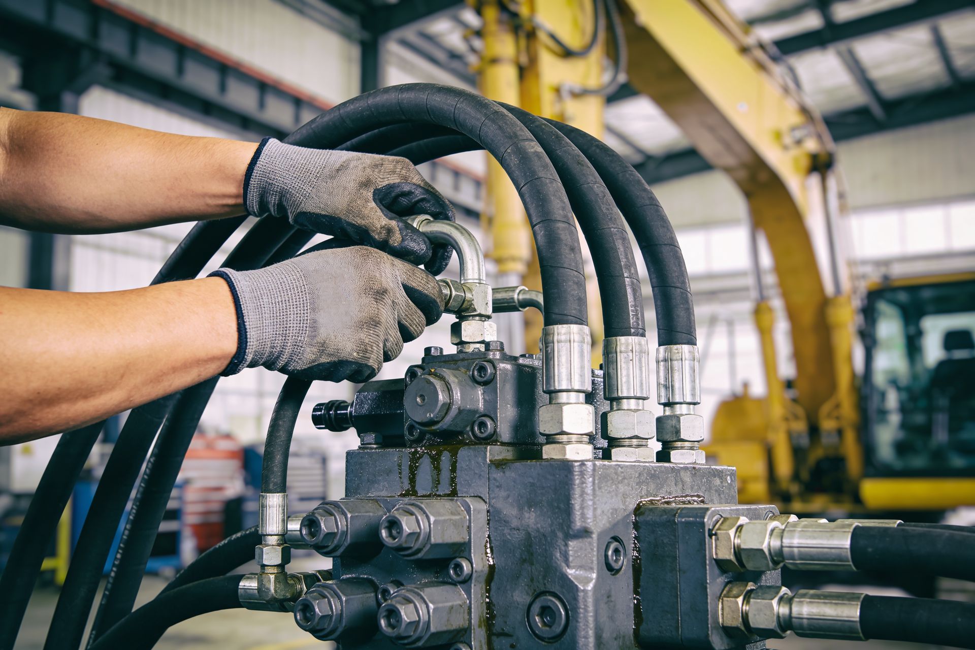 A mechanic's hands in gloves are connecting hydraulic hoses to a heavy machinery valve block.
