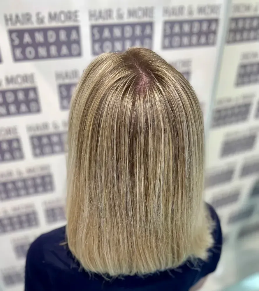 Blondes, glattes Haar mit helleren Strähnen. Im Hintergrund ist ein Salonlogo zu sehen.