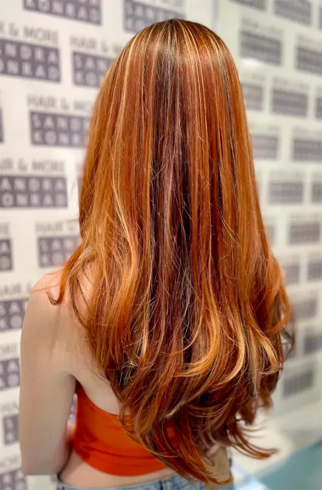 Frau mit langem, gewelltem, kupferfarbenem Haar mit Strähnchen und einem rostfarbenen Oberteil.