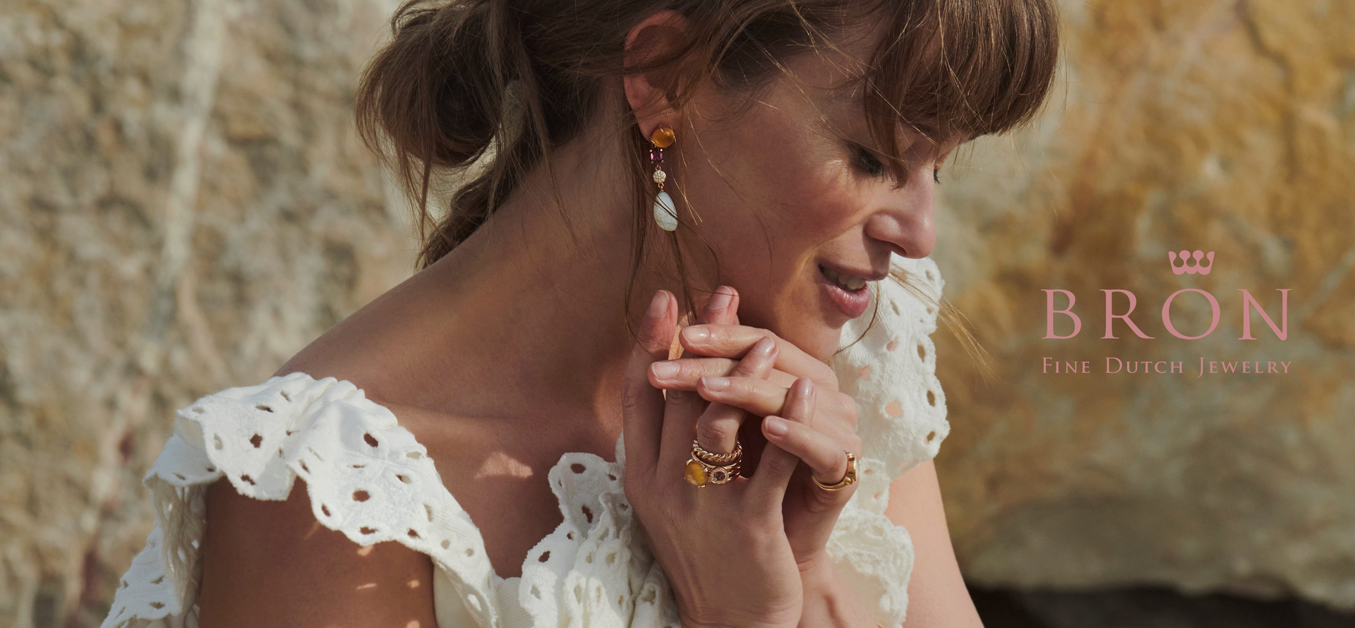 Een vrouw in een witte top met ajourwerk draagt ​​gouden oorbellen en ringen, haar handen gevouwen bij haar gezicht, bij een rotswand. Logo: BRON.