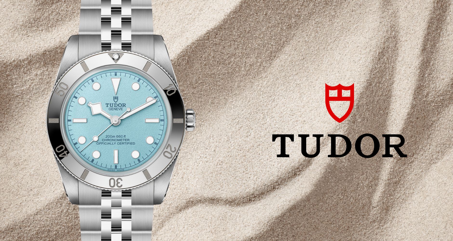 Zilveren Tudor-horloge met turquoise wijzerplaat op een beige stoffen achtergrond, Tudor-logo rechts.