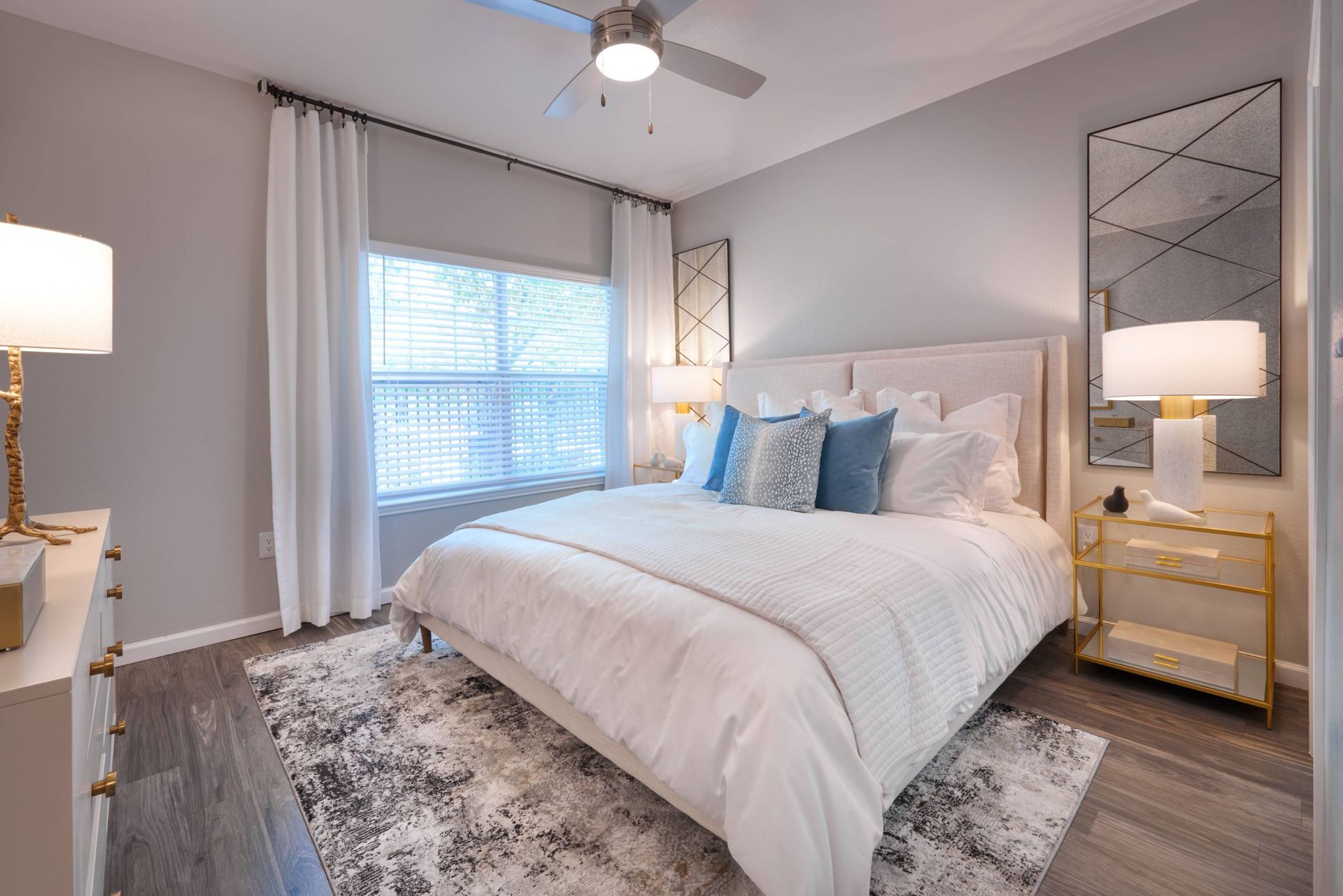 Bedroom with white bedding, beige headboard, two gold nightstands, lamps, ceiling fan, and large window.